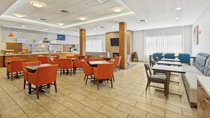 A hotel lobby with multiple dining tables and chairs, a reception desk, and a television screen.