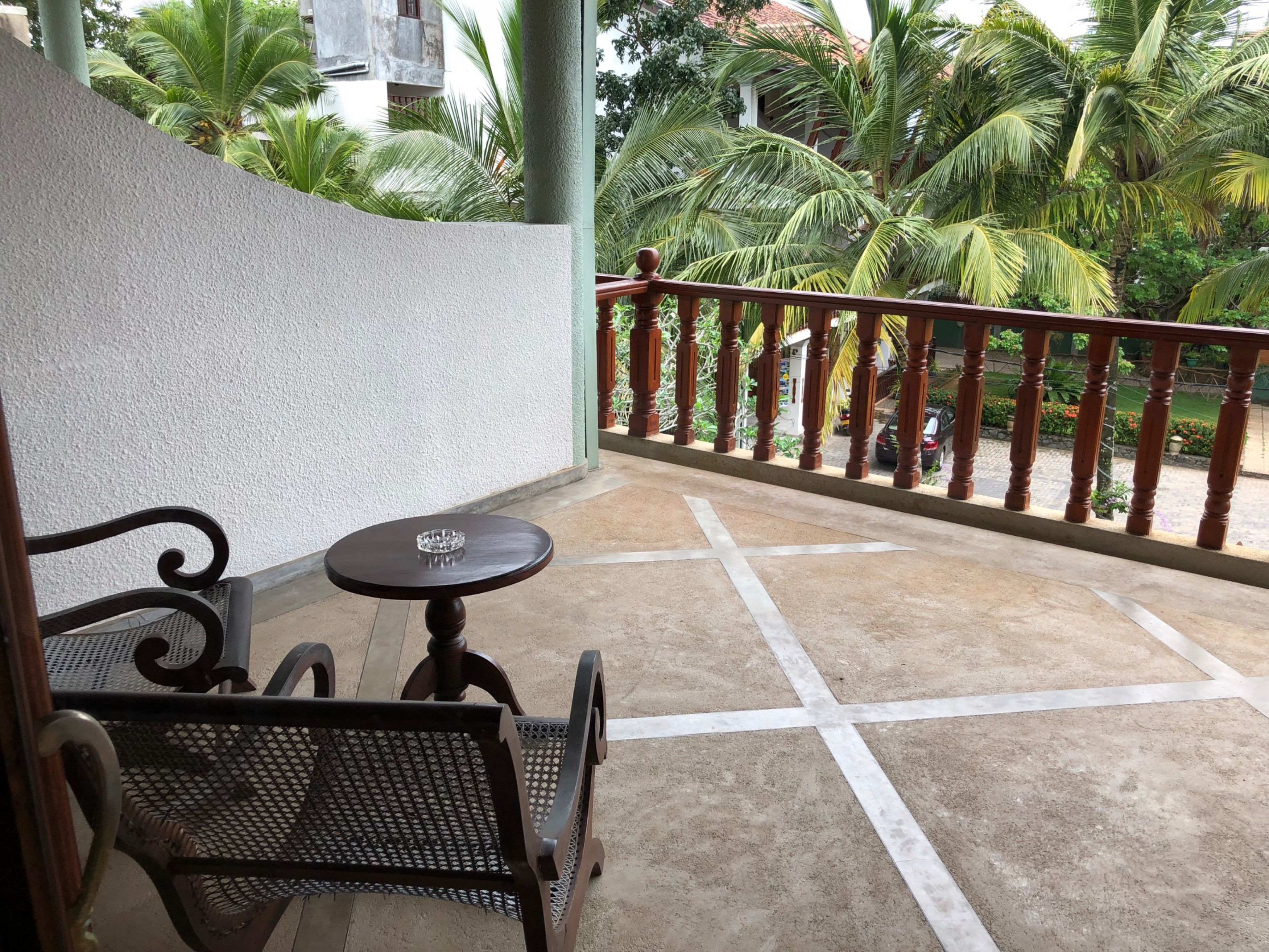 A balcony with a table and chairs, a railing, and a view of palm trees and a parked car.