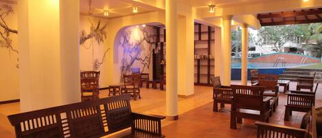A spacious lobby with wooden furniture, a marble wall, and a pool area visible through an archway.