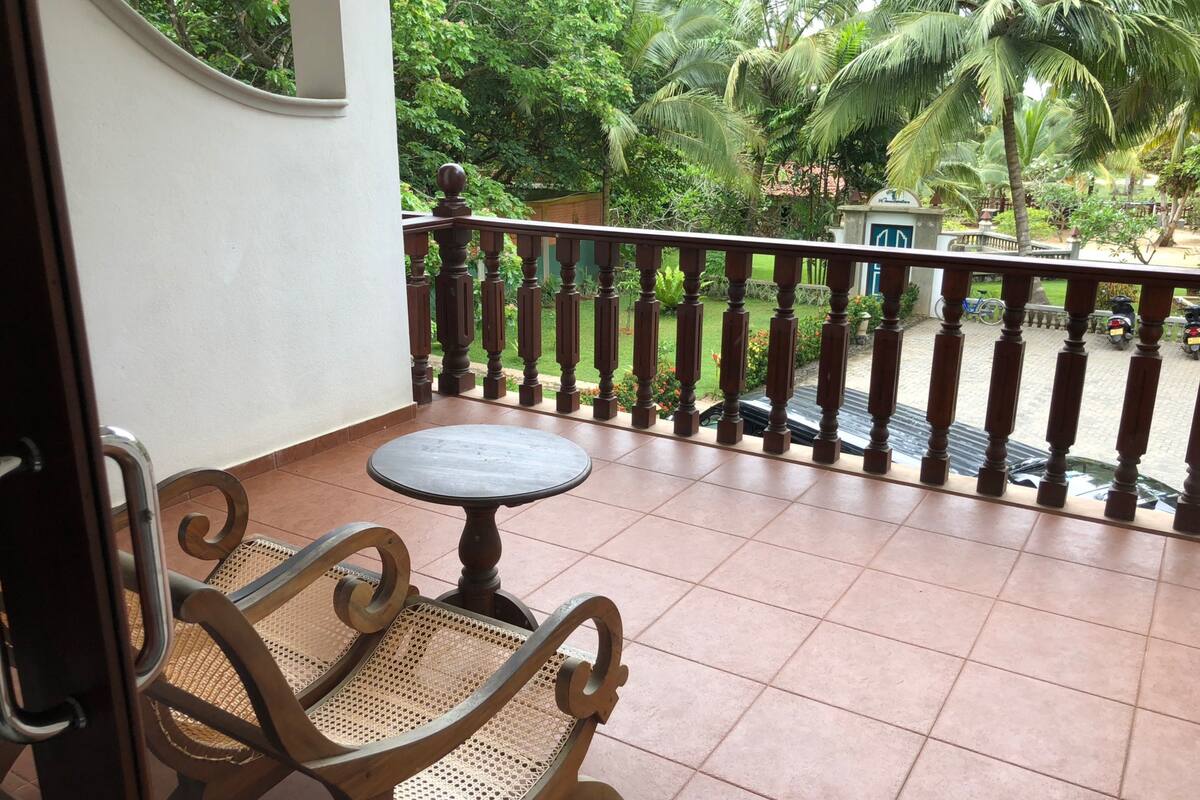 A balcony with a round table and two wicker chairs, overlooking a garden with palm trees and a paved area with parked scooters.