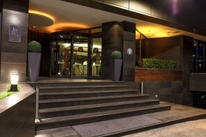 Entrada moderna de hotel com porta de vidro, degraus de acesso e um vaso de planta ao lado da porta.
