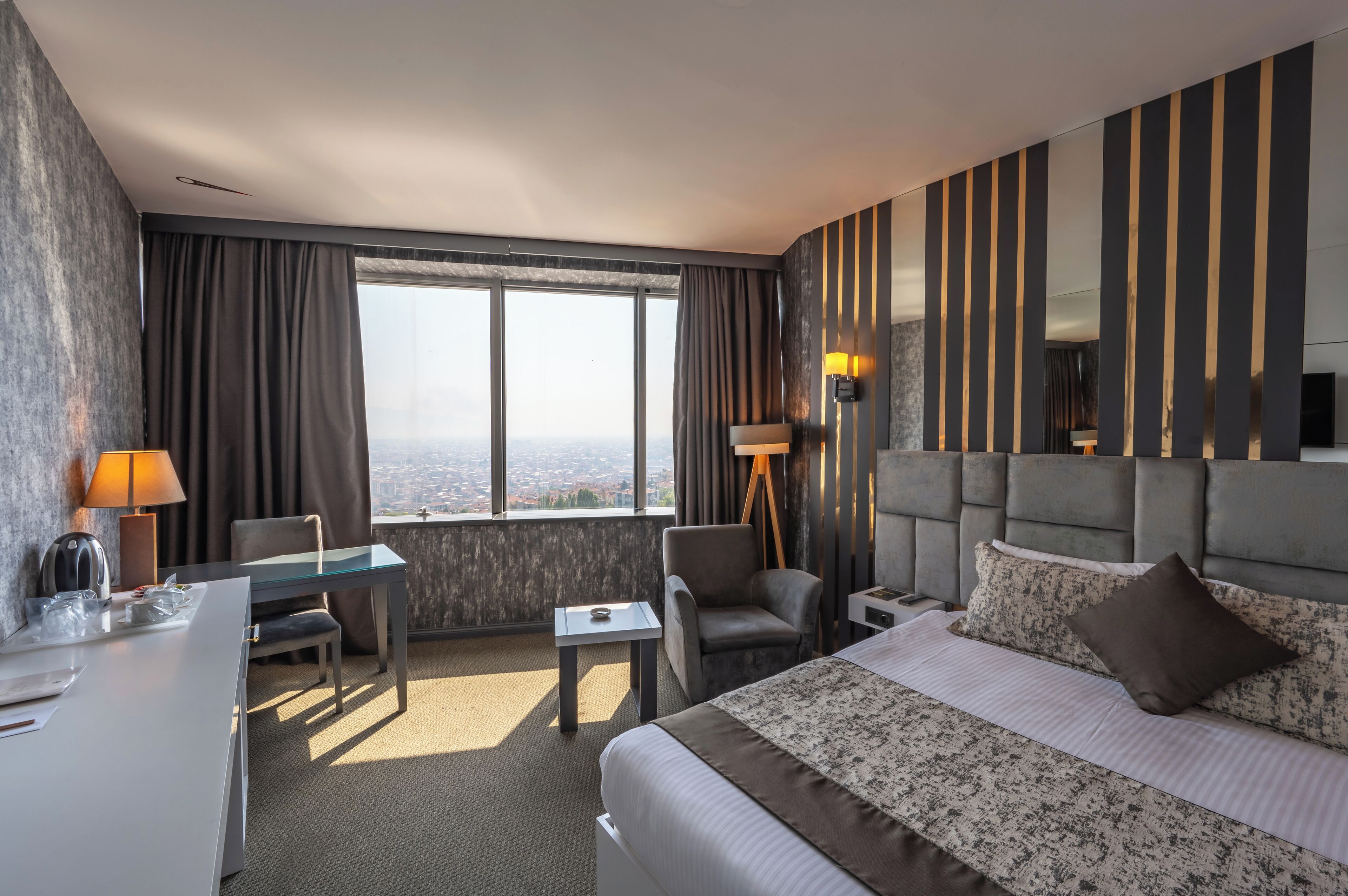 Quarto de hotel com uma cama grande, uma escrivaninha, uma cadeira, uma mesinha e vista da cidade por grandes janelas.