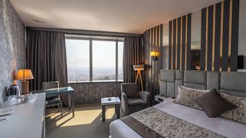 Quarto de hotel com uma cama grande, uma escrivaninha, uma cadeira, uma mesinha e vista da cidade por grandes janelas.