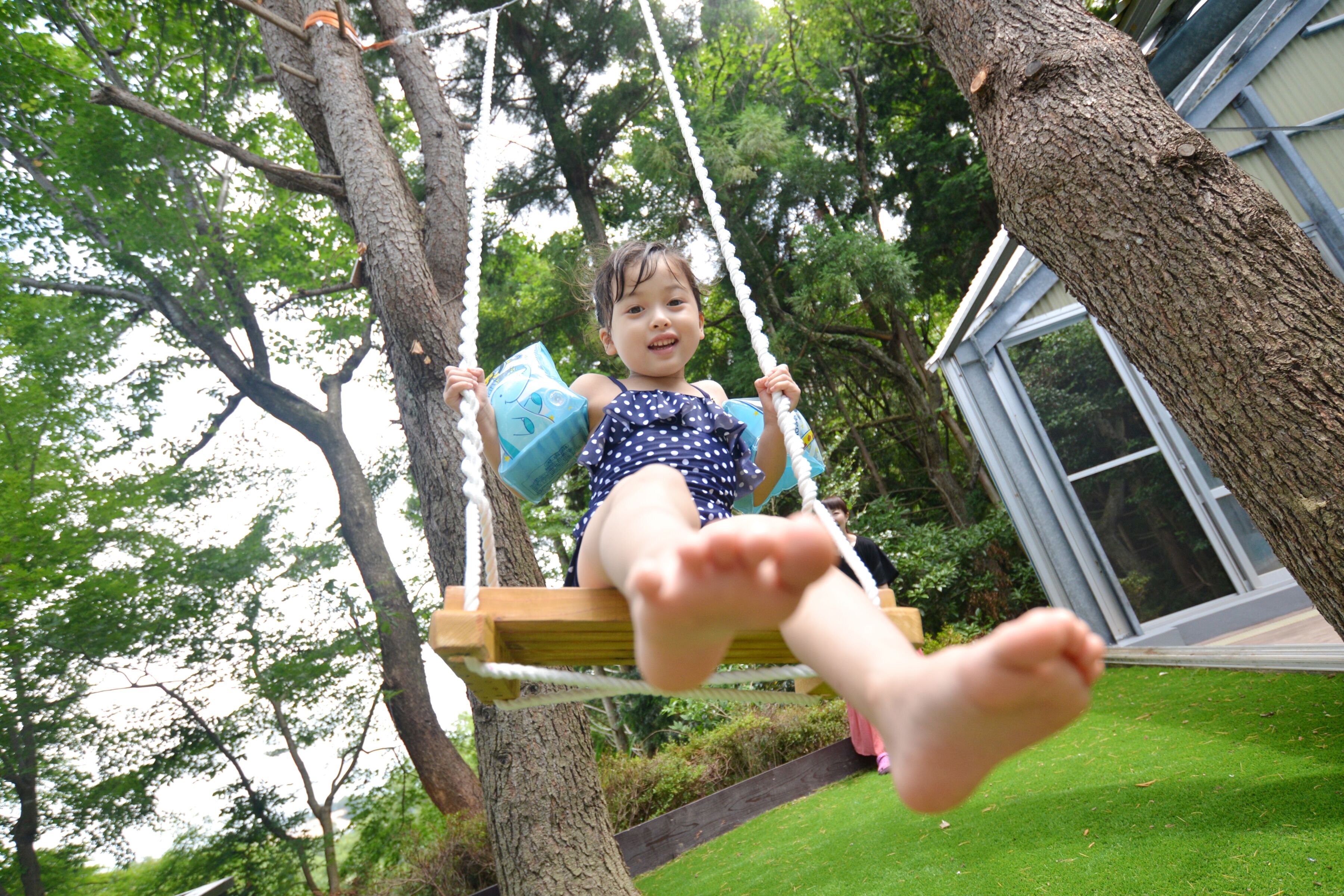 A child on a swing in a lush, green outdoor setting with trees and a building in the background.