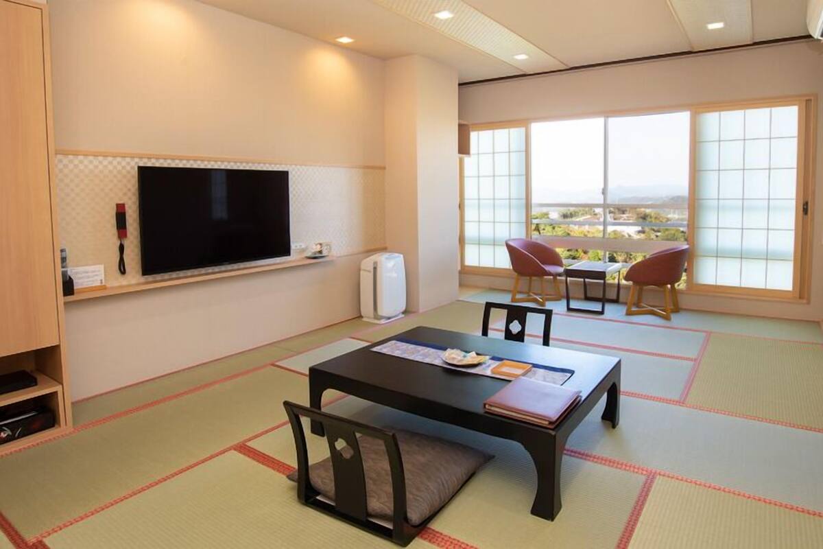 A room with tatami mats, a television, a table with chairs, and a view through large windows.