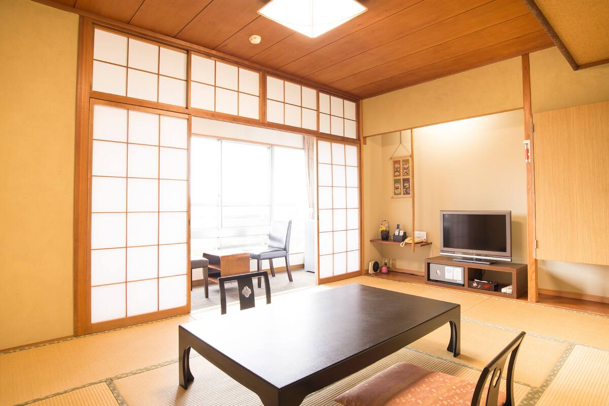A traditional Japanese room with tatami flooring, a low table, chairs, a television, and sliding doors.
