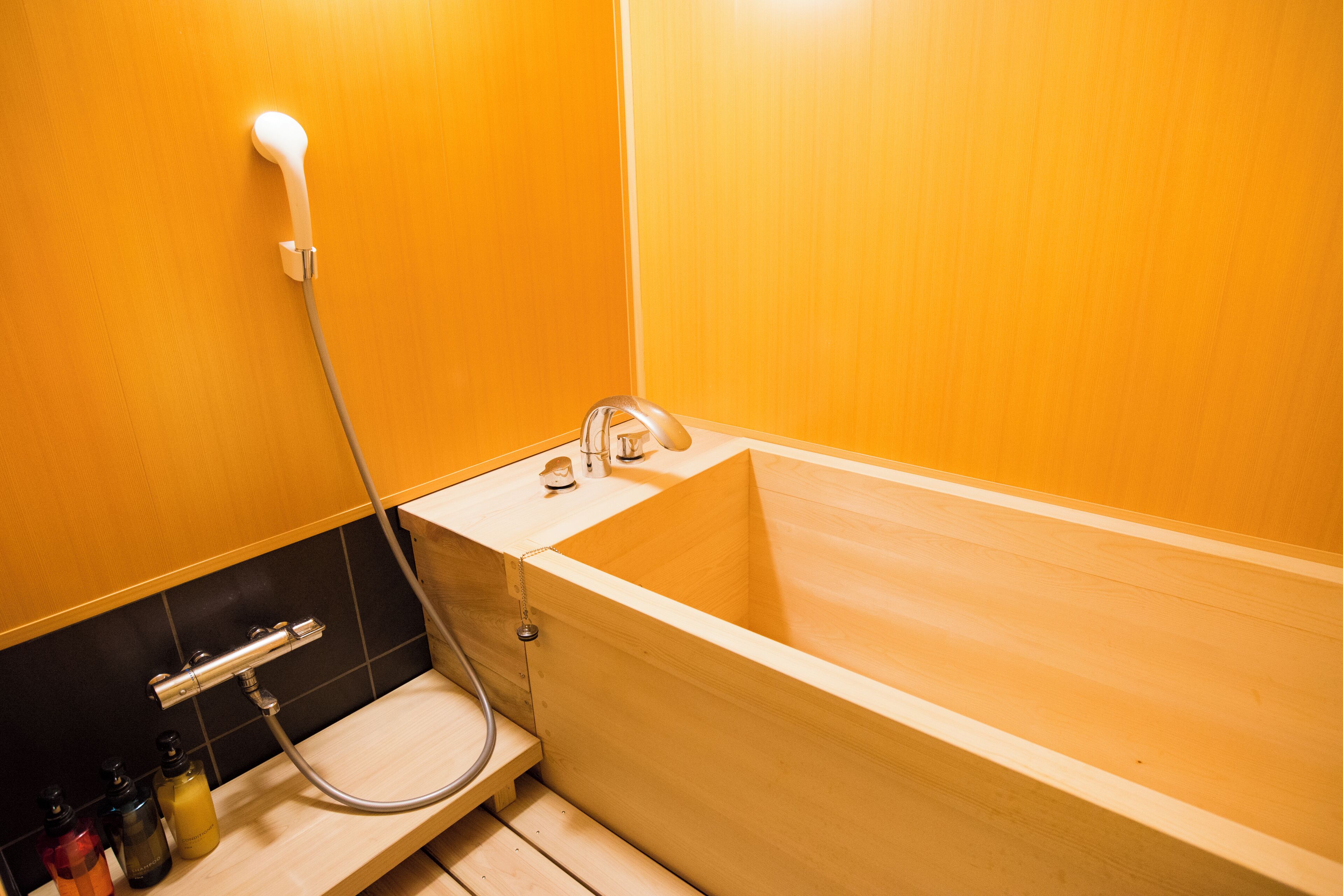 A wooden bathtub with a shower head and faucet, a wooden shelf with toiletries, and a black tiled wall.