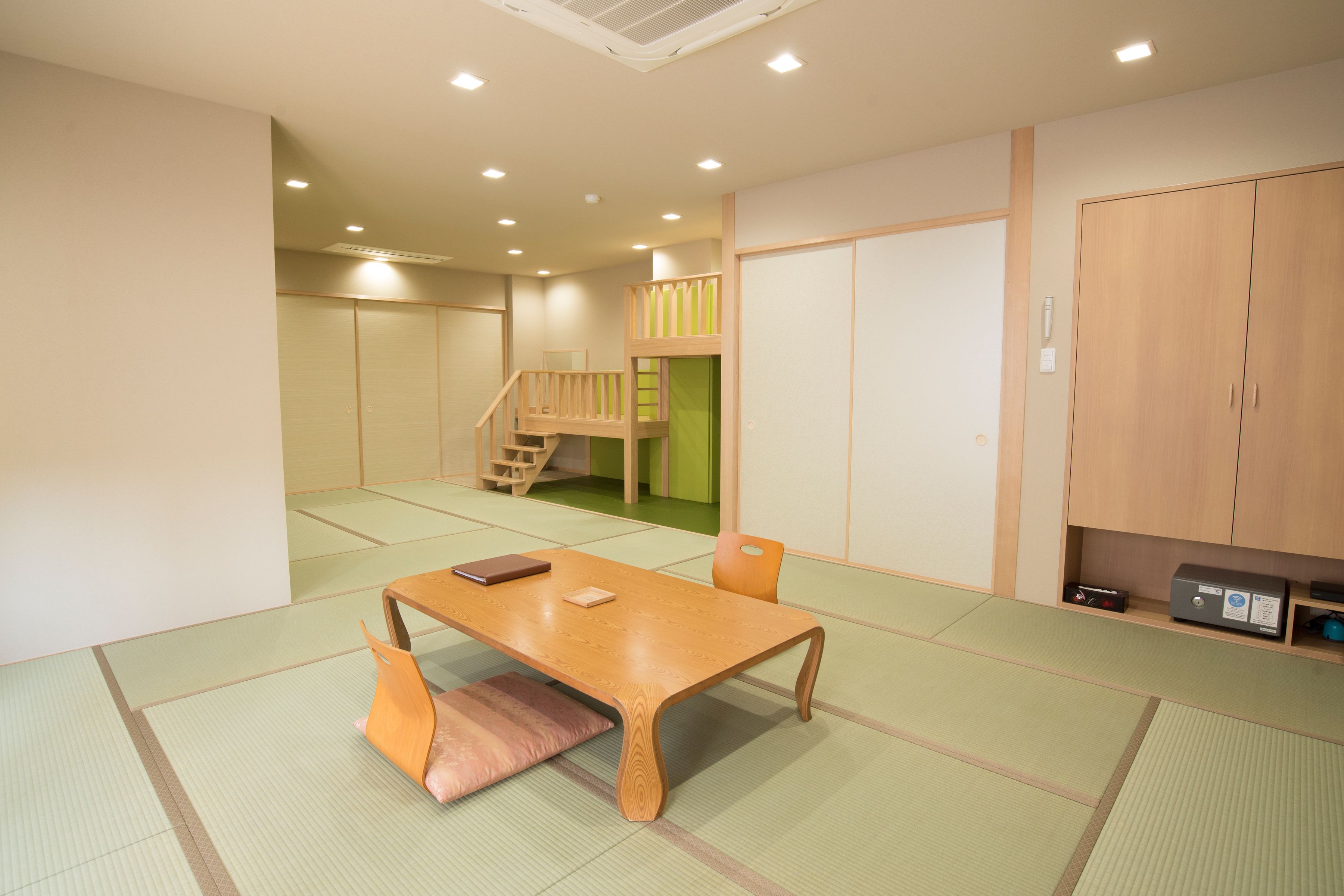 Una habitación japonesa tradicional con suelo de tatami, una mesa de madera baja, un banco acolchado, una puerta corredera verde, una escalera de madera y un armario.
