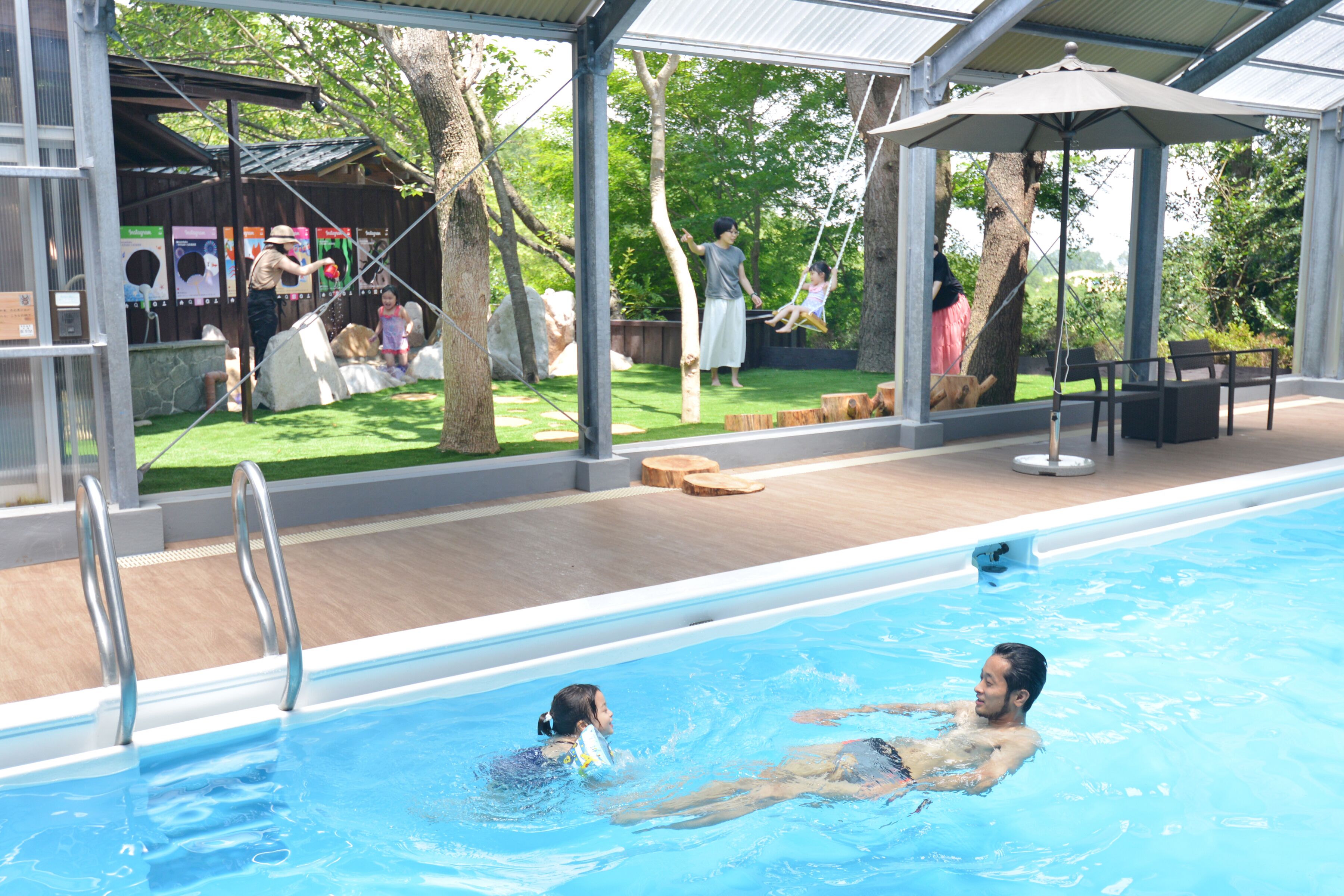 Piscina con área techada, terraza de madera y vista a un jardín con árboles y un columpio.