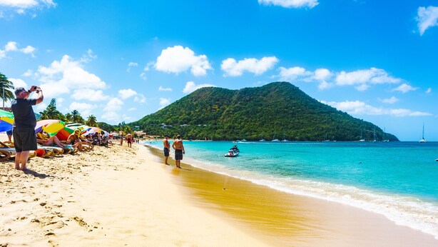 A beach with a clear sky, a mountain in the background, and a few people and boats in the water. - Cleopatra Villas (Gros Islet)