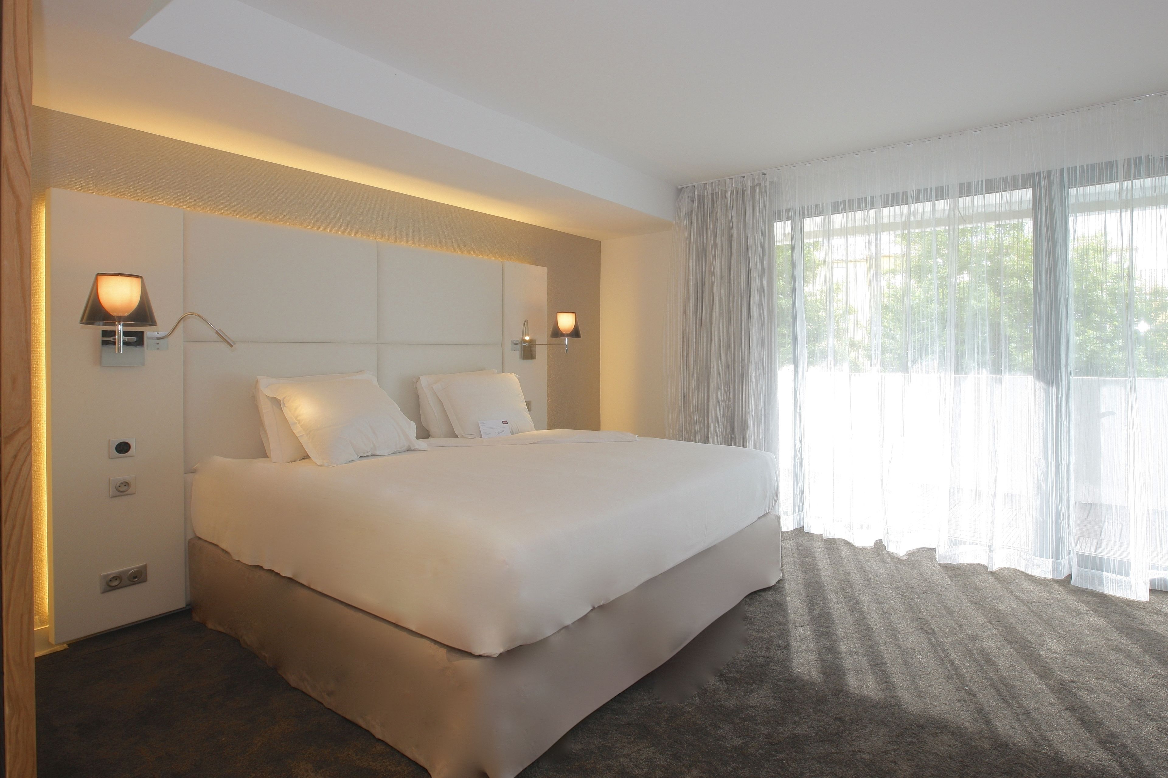 A large bed with white linens, two wall-mounted lamps, and sheer curtains on a window.