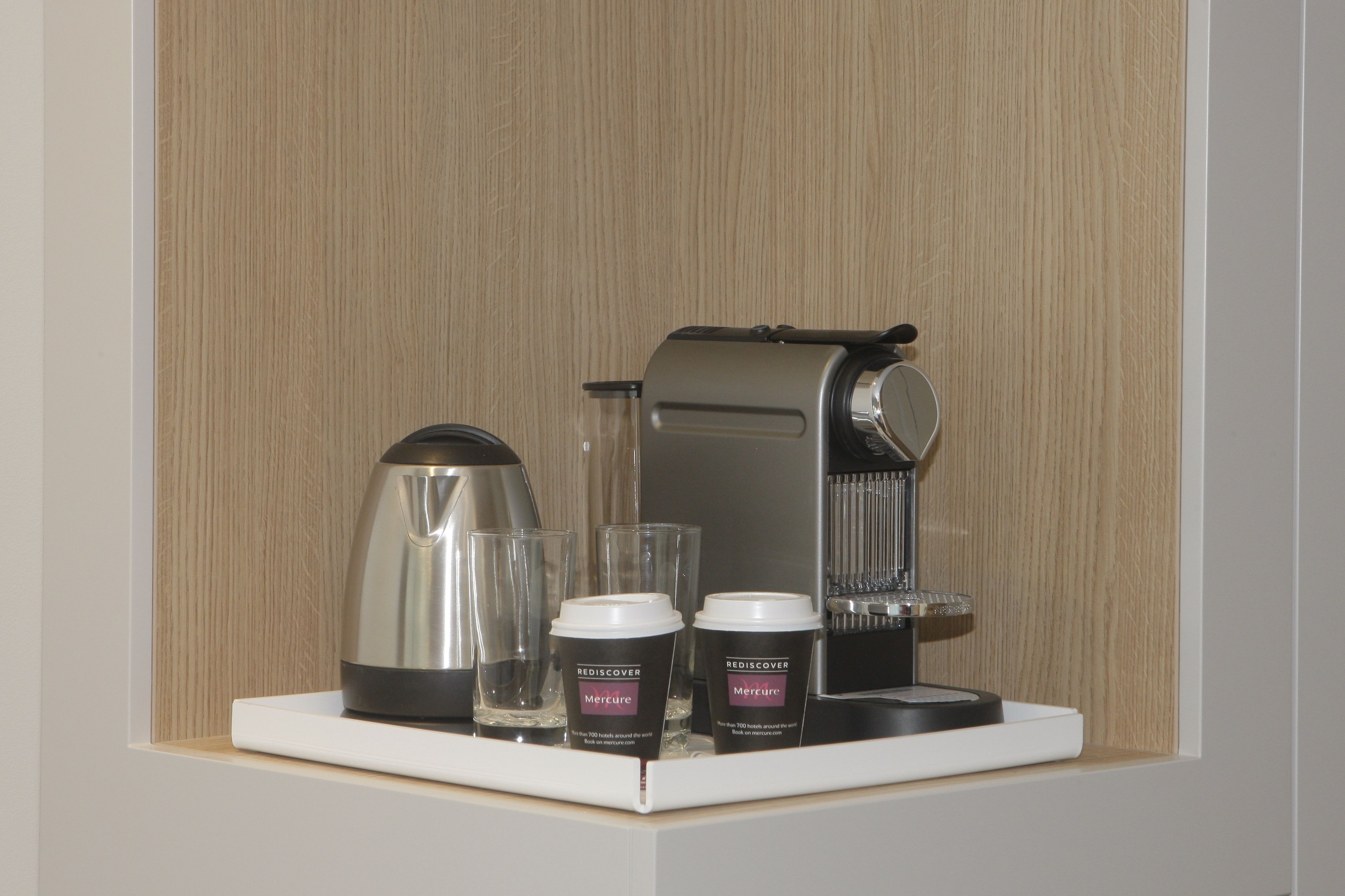 A coffee machine, two glasses, and two containers of meringue on a white shelf against a wooden wall.