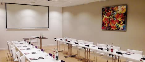 Un salón de conferencias con mesas y sillas blancas, y una pantalla para proyector. En la pared, un cuadro abstracto de gran tamaño.