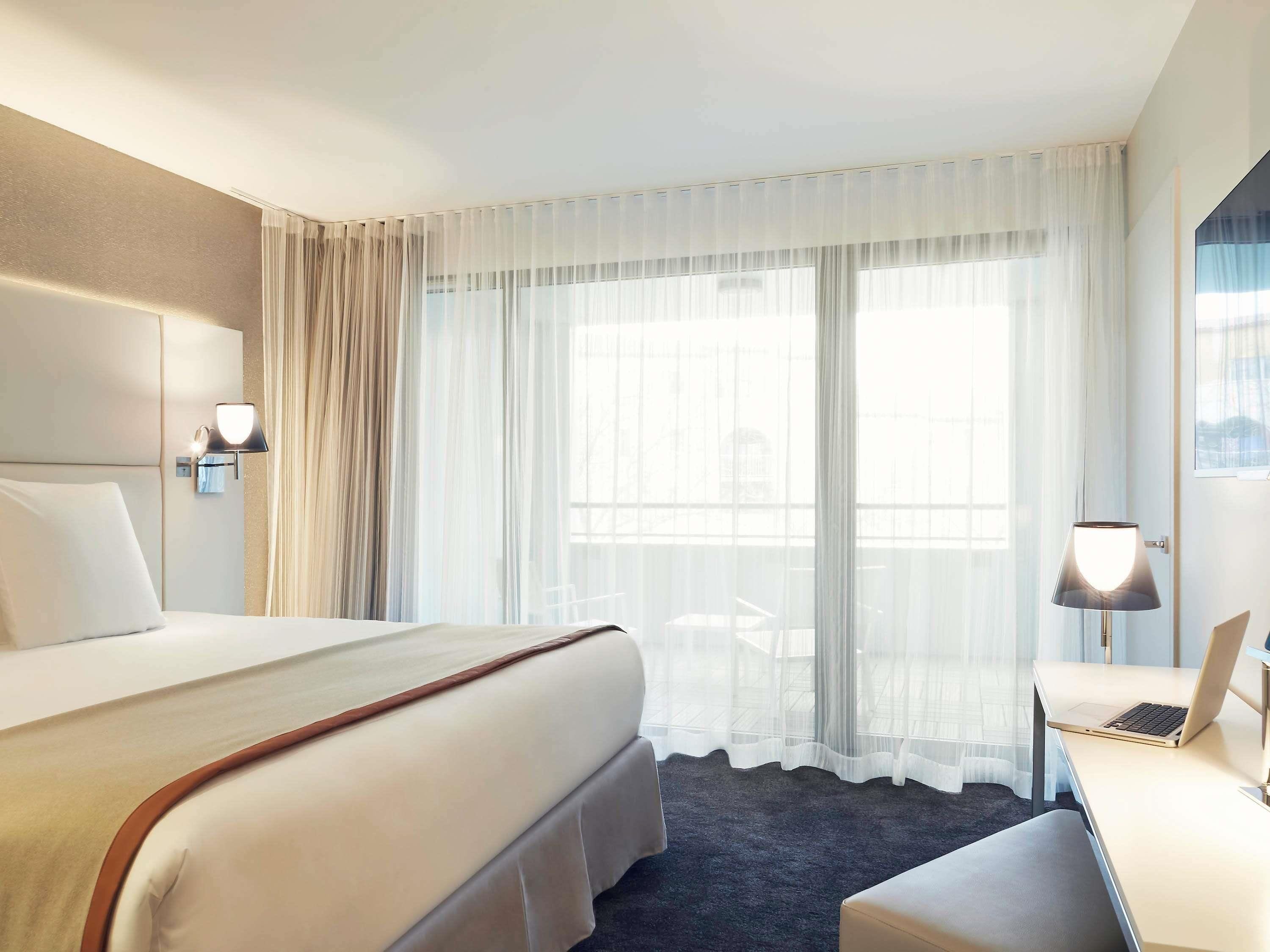 Une chambre d’hôtel avec un grand lit, un bureau avec un ordinateur portable, une chaise et une fenêtre avec des rideaux légers.