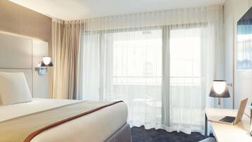 Une chambre d’hôtel avec un grand lit, un bureau avec un ordinateur portable, une chaise et une fenêtre avec des rideaux légers.