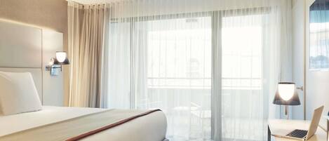 A hotel room with a large bed, a desk with a laptop, a chair, and a window with sheer curtains.