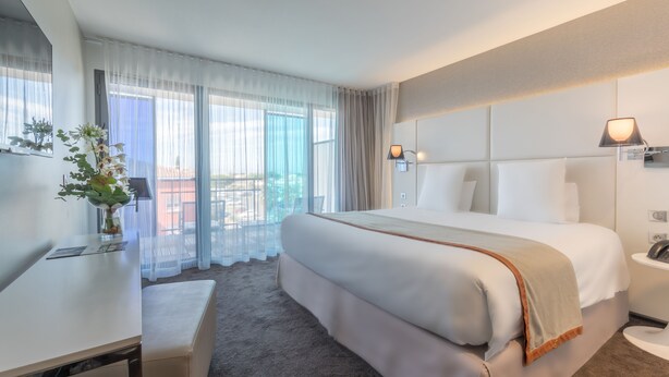 A modern hotel room with a large bed, a desk, and a view of the city through sheer curtains. - Mercure Golf Cap d'Agde (Agde)