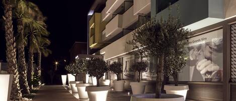 Vista nocturna de un edificio moderno con plantas en macetas iluminadas y un letrero que dice 'Des Grands Vins'.