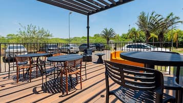 Un patio al aire libre con mesas y sillas, una pérgola y vistas a coches aparcados y follaje.