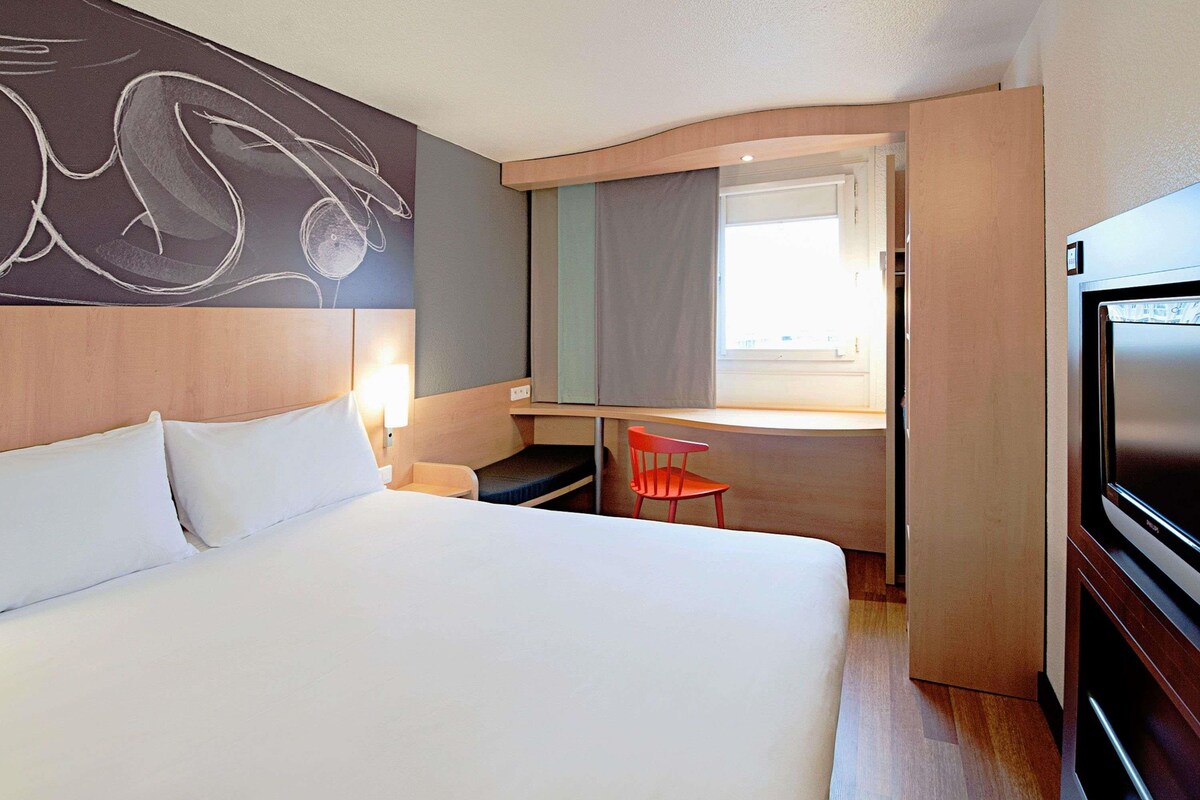 Ein Hotelzimmer mit Bett, Schreibtisch, Sessel, Fernseher und Fenster.