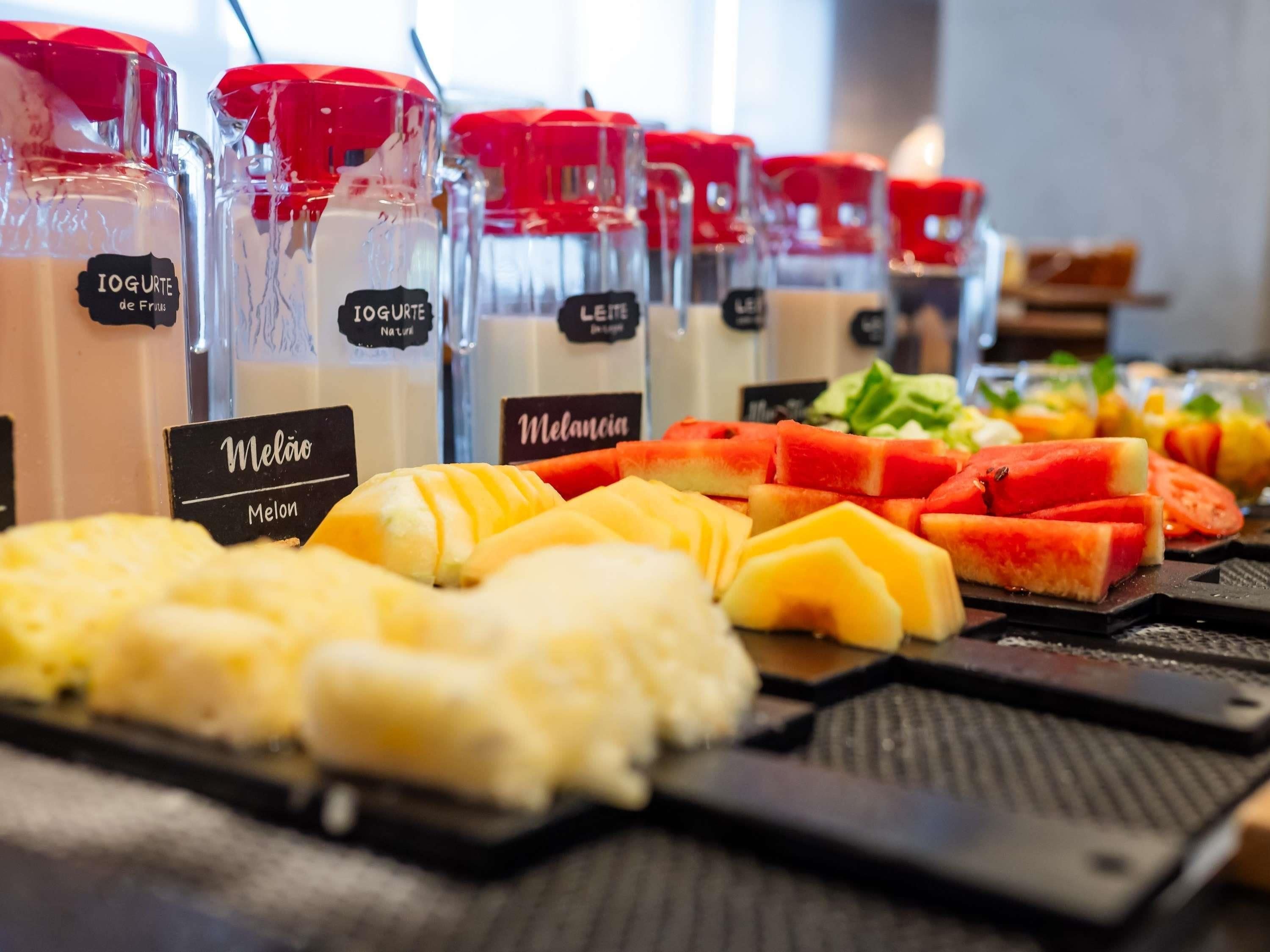 Variedad de bebidas de yogur en recipientes transparentes con tapas rojas, junto con frutas frescas como melón y sandía.