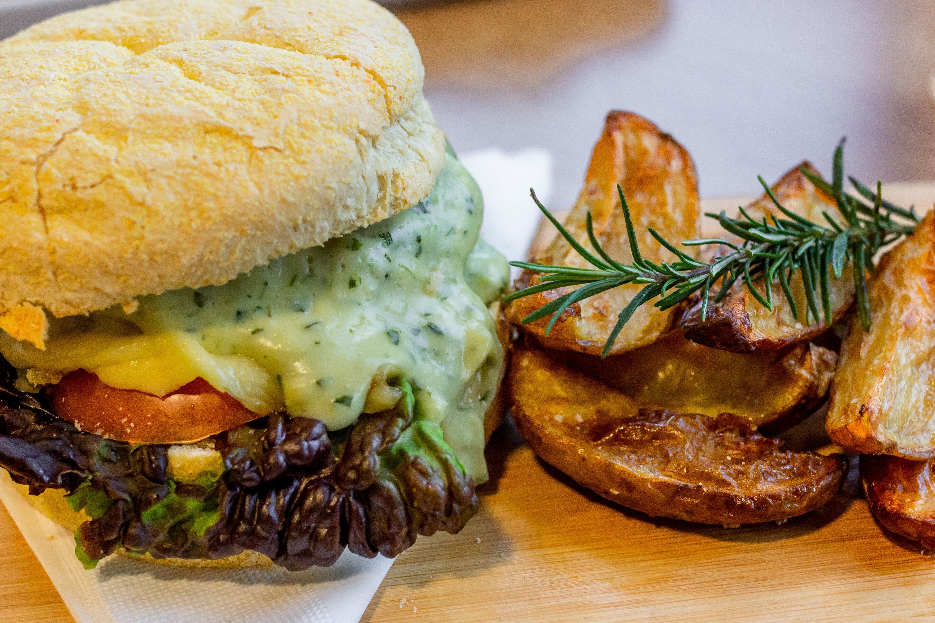 A sandwich with a creamy sauce, lettuce, tomato, and cheese, accompanied by roasted potatoes and a sprig of rosemary.