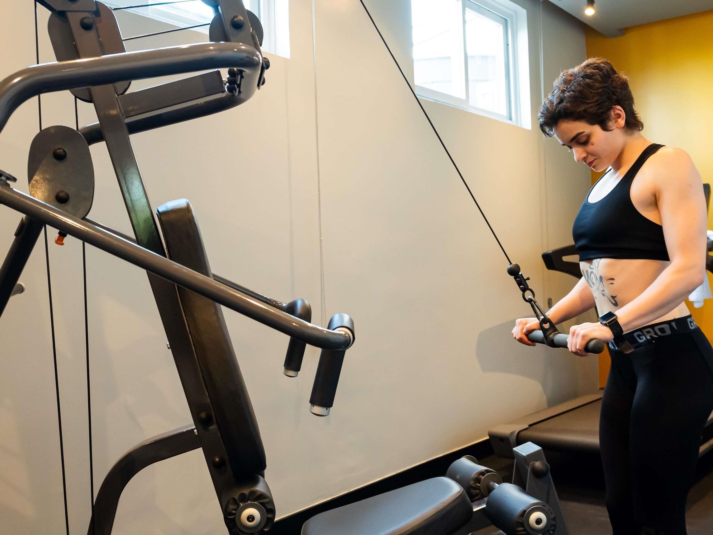 A gym with exercise equipment, a person using a cable machine, and a window with natural light.