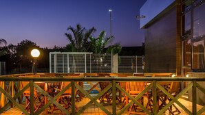 An outdoor dining area with wooden chairs and tables, a metal fence, and a building with large windows in the background. - ibis Porto Alegre Assis Brasil (Porto Alegre)
