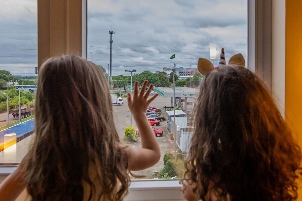 Two children looking out of a window with a view of a parking lot and buildings.