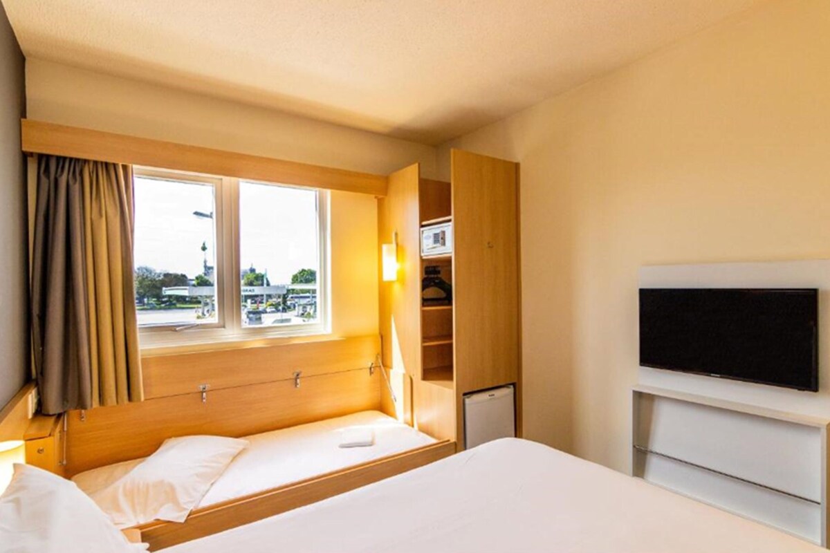 A hotel room with a bed, a TV, a window with curtains, and a view of the outside.