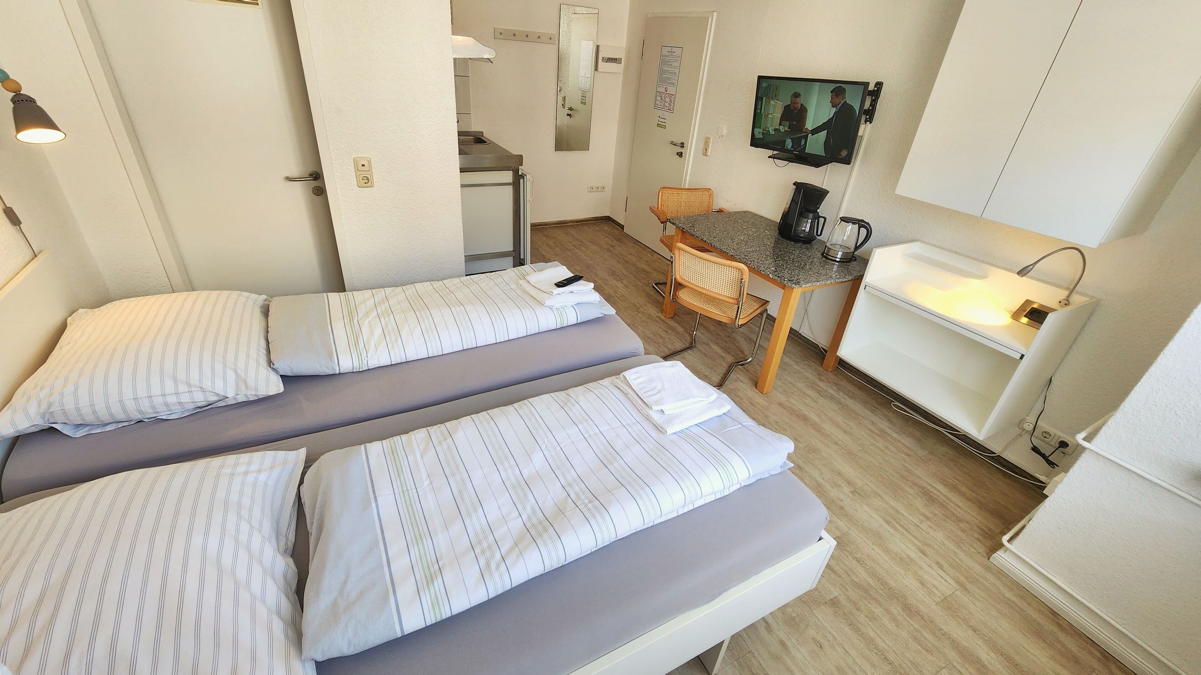 A compact hotel room with two beds, a small table, a chair, a TV, and a coffee machine.