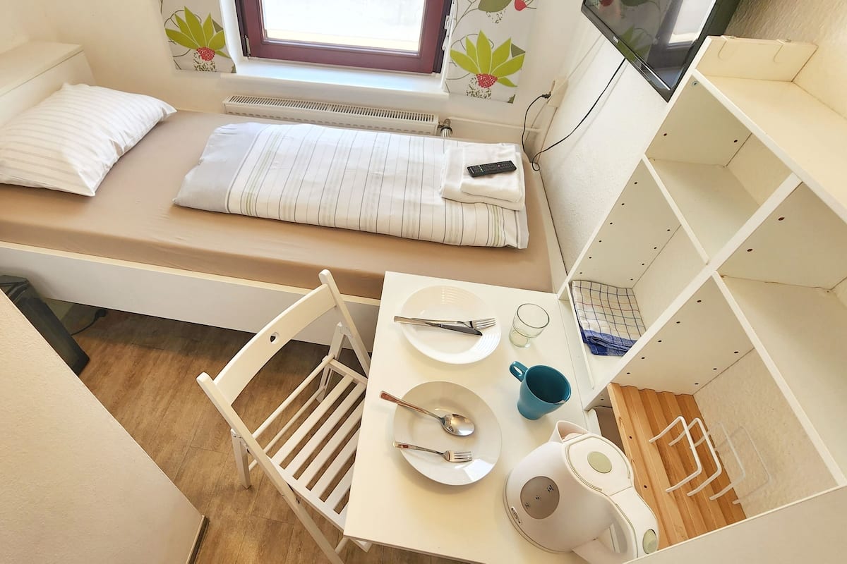 A compact room with a bed, a small table with a kettle, a chair, and a window with a view.
