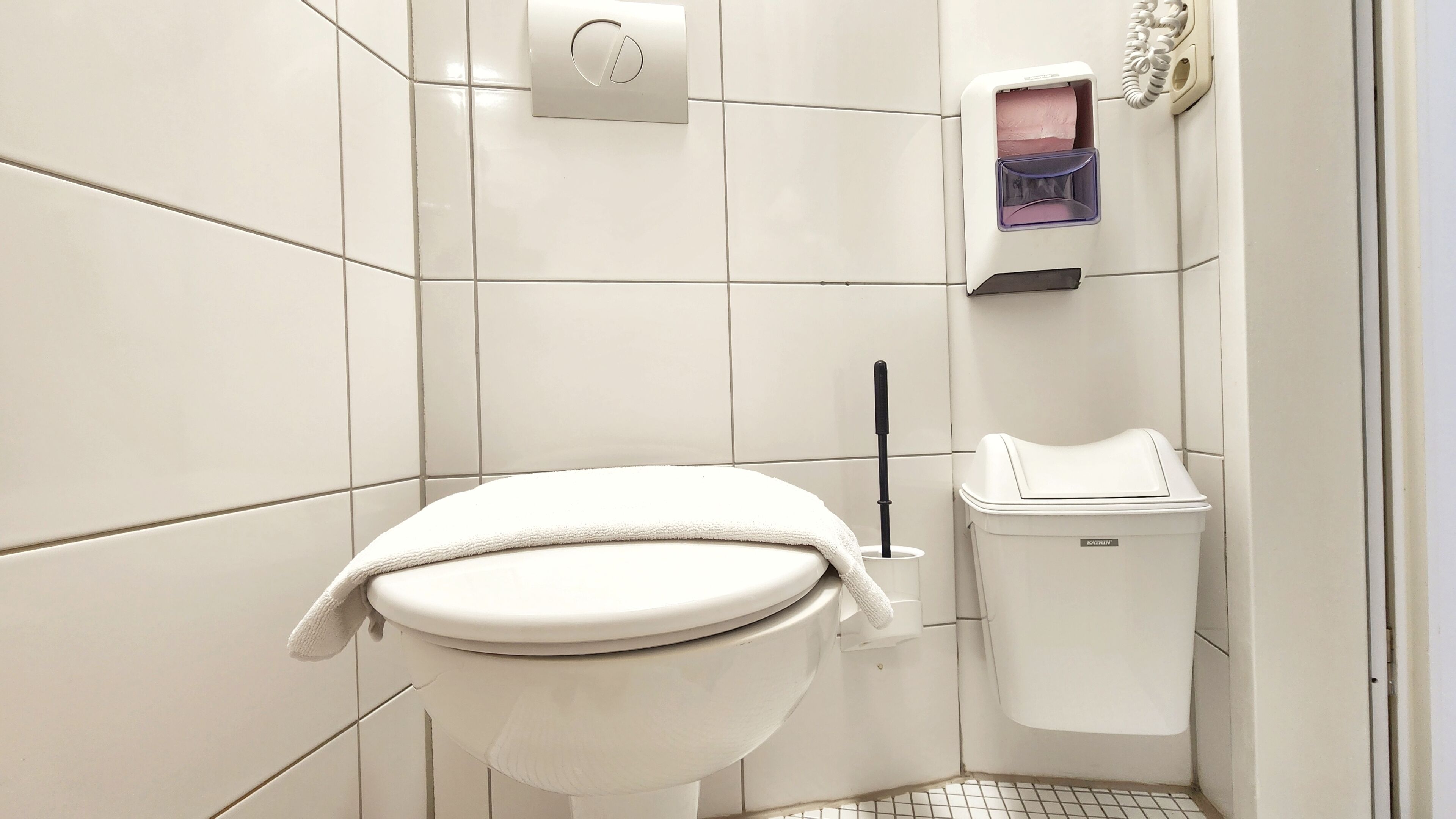 A white toilet with a towel on the seat, a toilet paper holder, and a trash bin in a corner bathroom with white tiled walls.