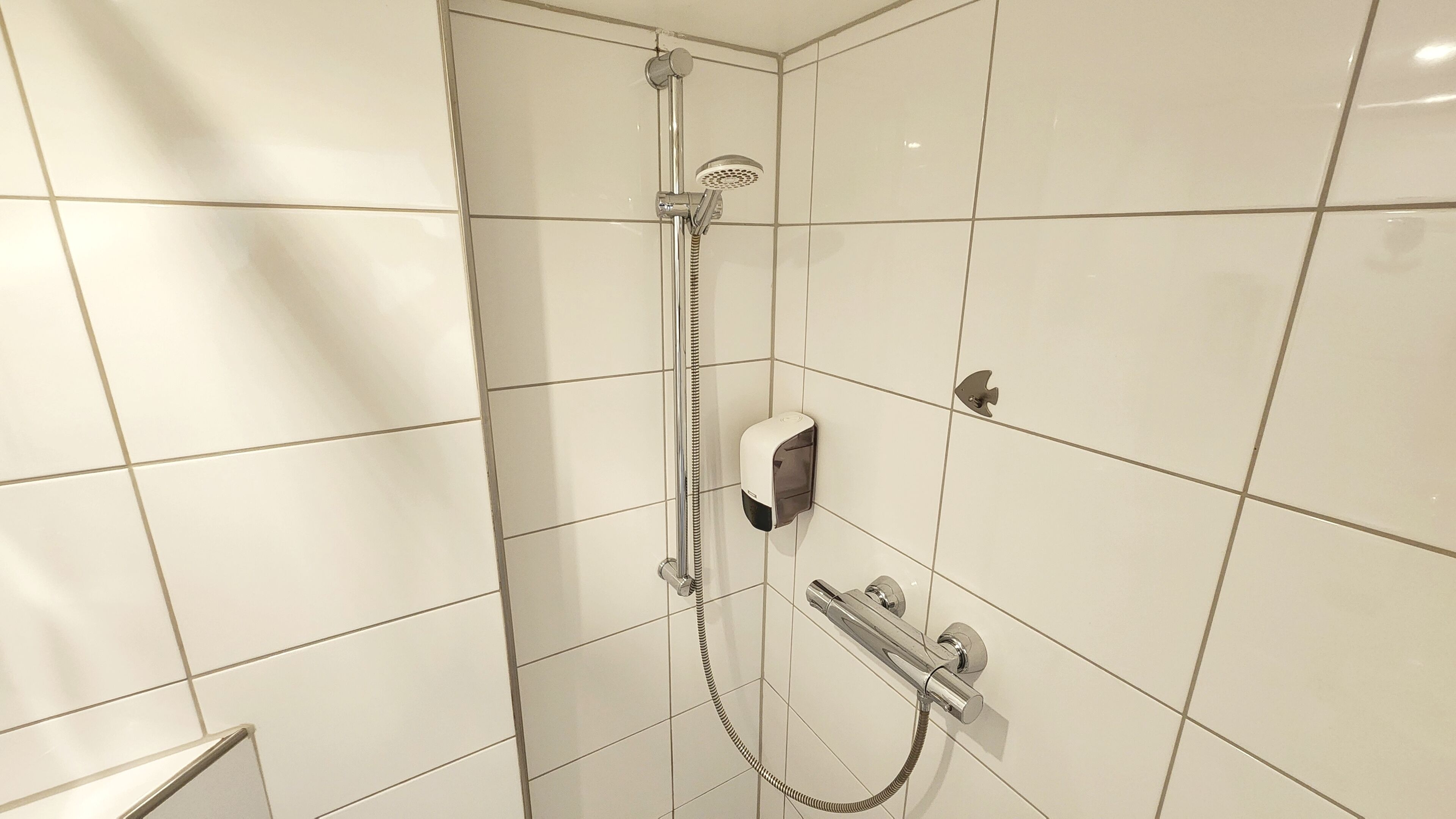 Un baño con paredes de azulejos blancos, una ducha con cabezal de ducha manual, un dispensador de jabón manual y un perchero para toallas.