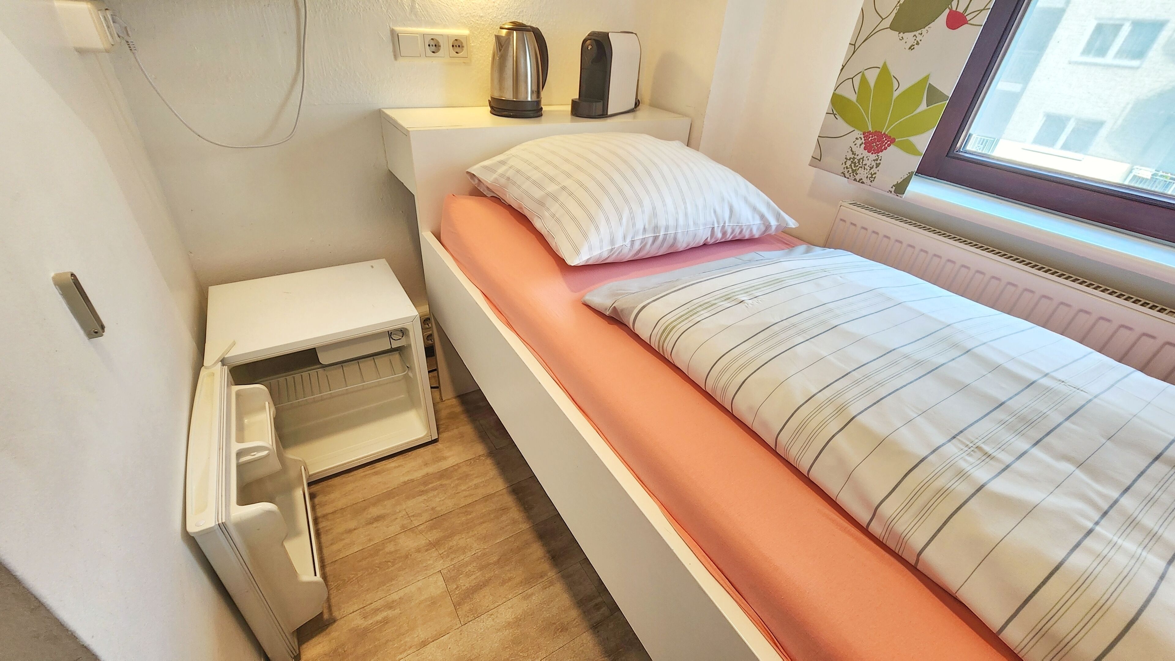 Habitación de hotel compacta con cama, mesita de noche pequeña, calefacción integrada y ventana con vistas a edificios.