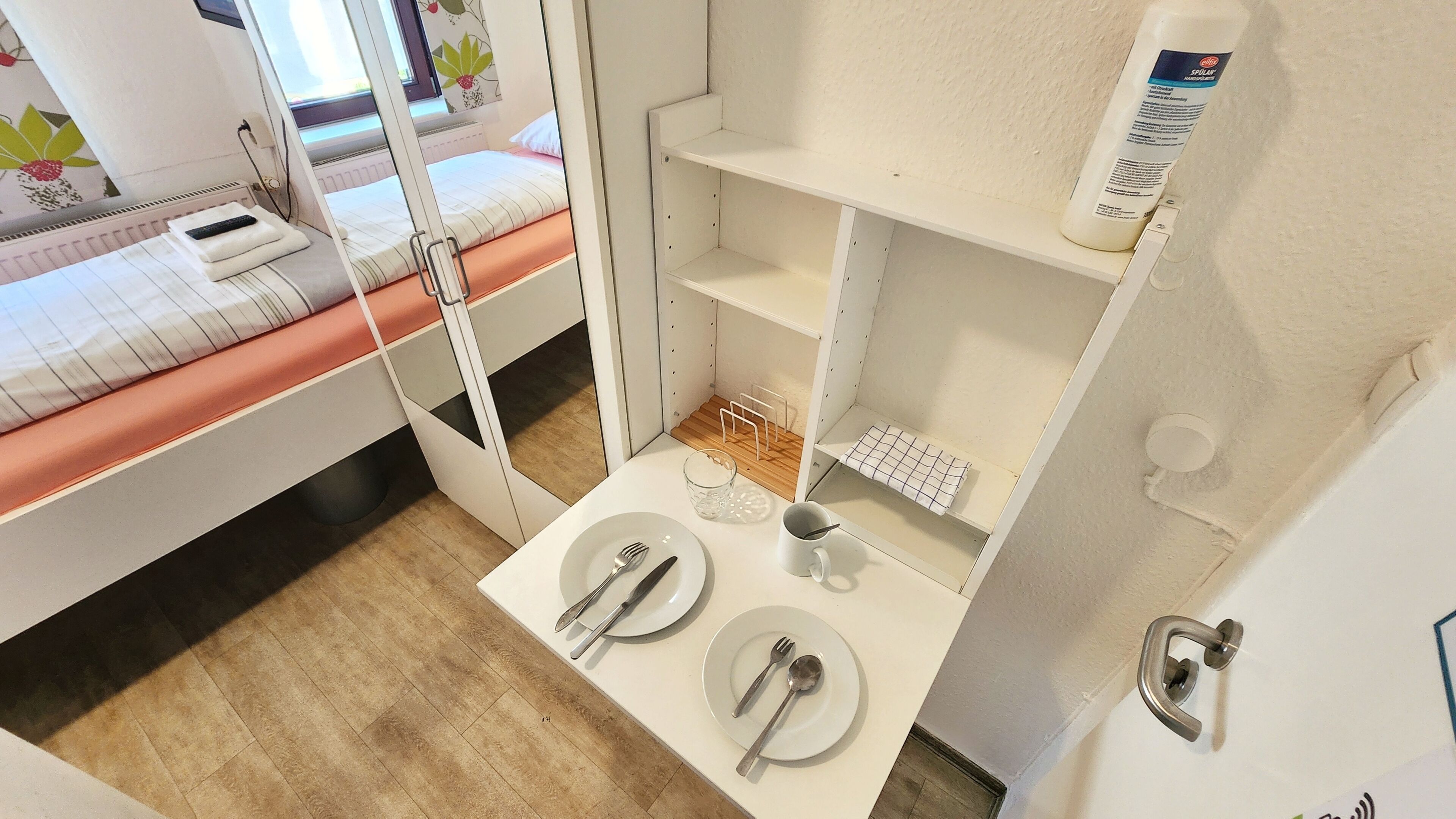 Una habitación de hotel pequeña con una cama, una mesita con una taza y cubiertos, y un estante con una botella.