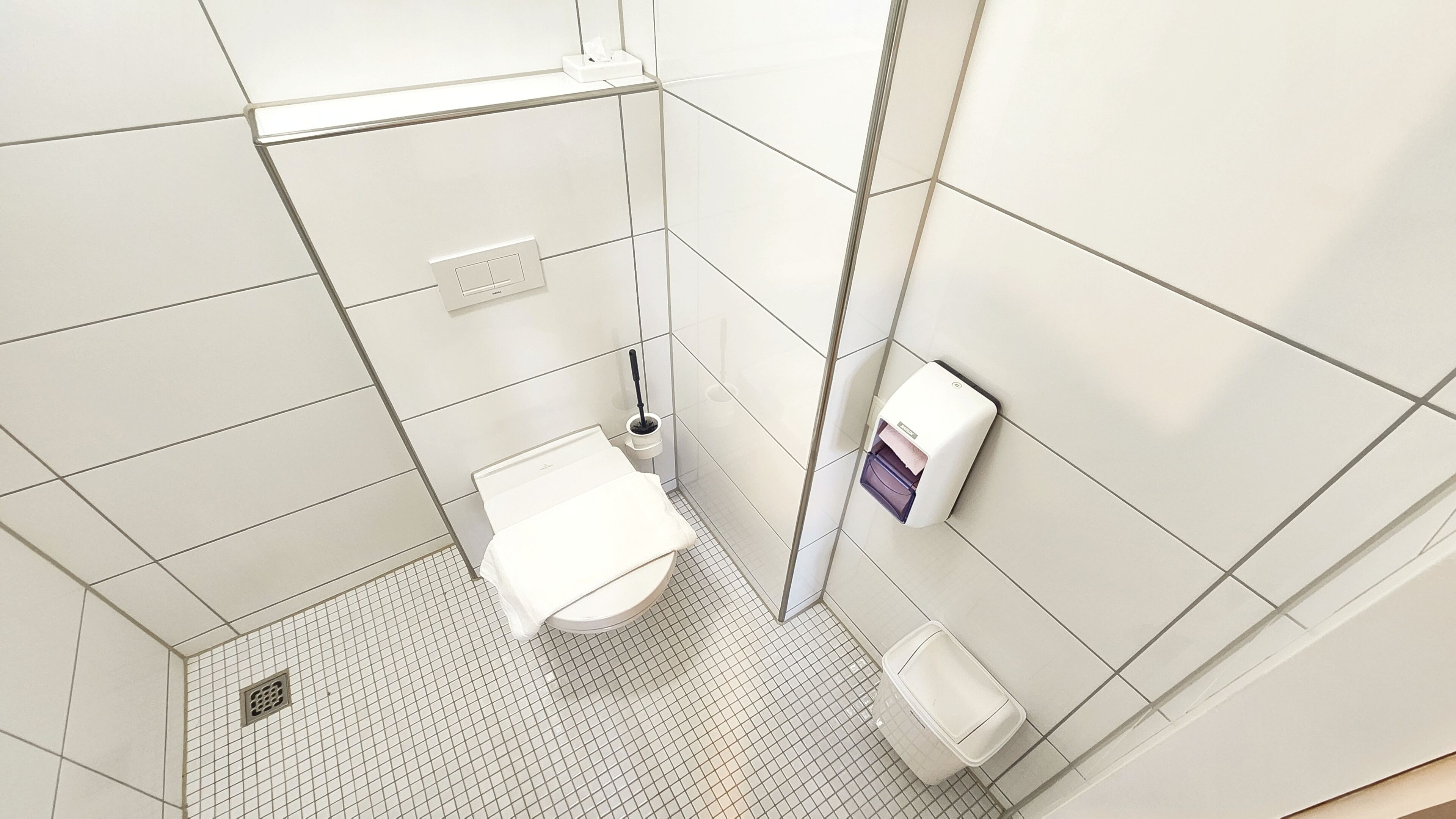 A small, modern bathroom with white tiles, a toilet, a hand dryer, and a trash bin.