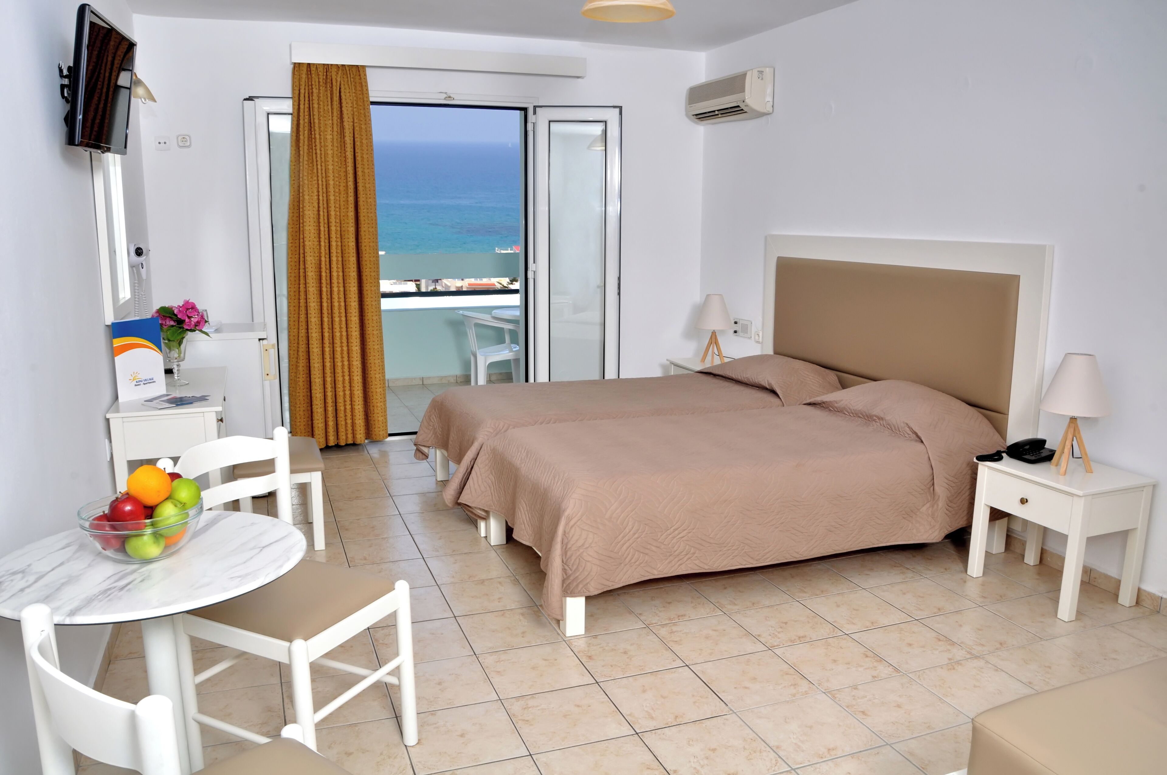 A hotel room with a bed, a desk with a chair, a small table with fruit, a TV, and a balcony with a view of the sea.