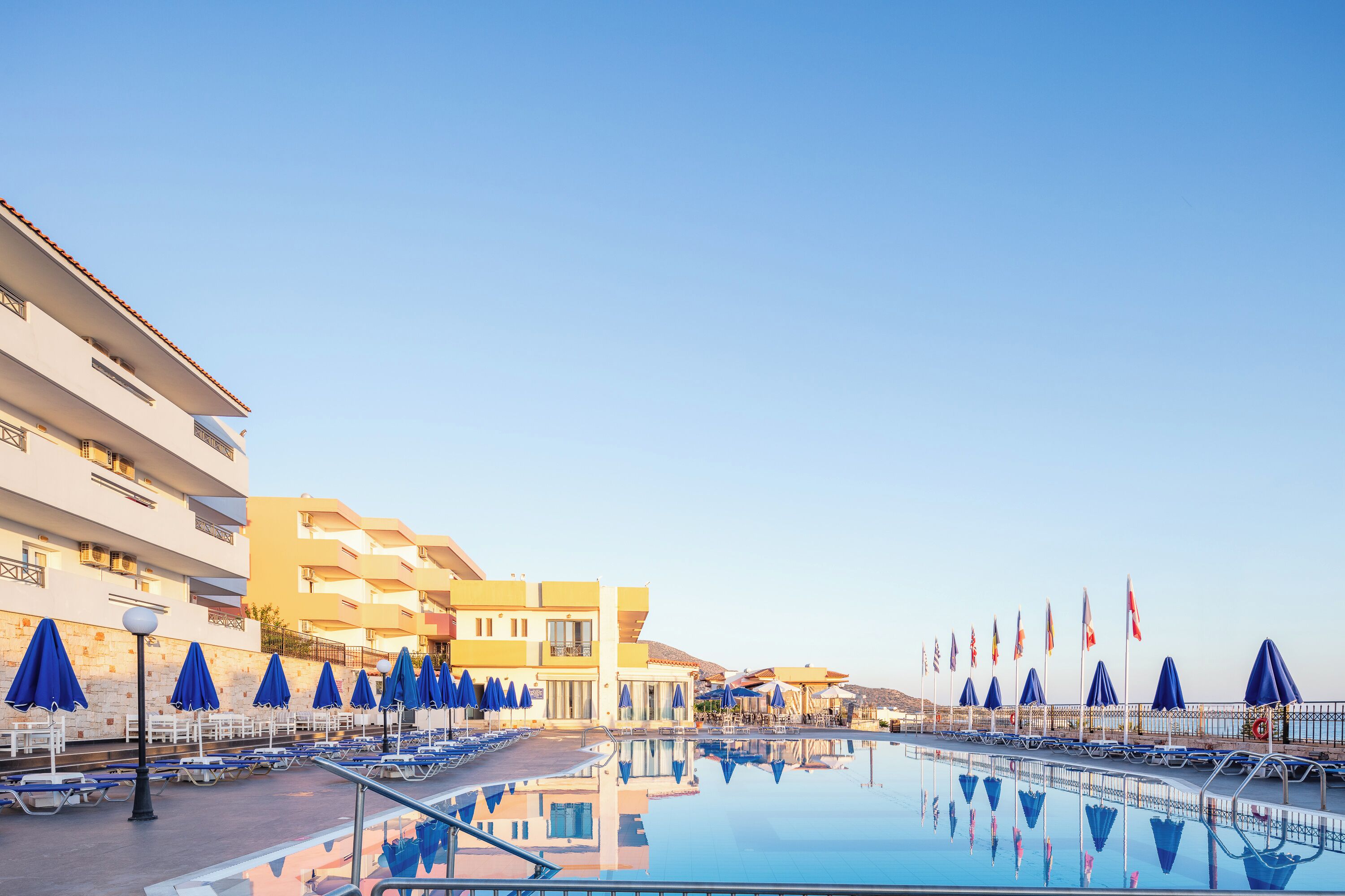 Zona de piscina de hotel con tumbonas y sombrillas, reflejando la piscina y los edificios.