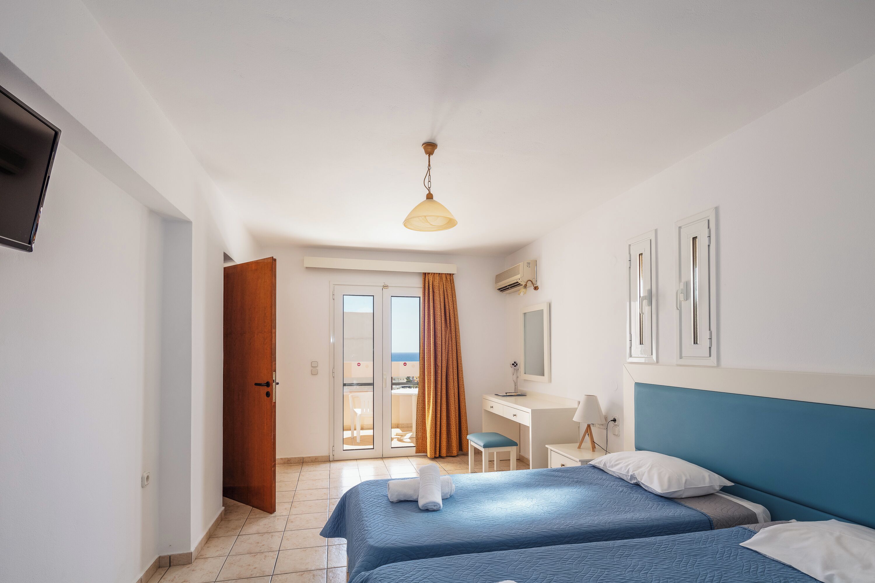 A hotel room with two beds, a desk, a chair, a TV, and a balcony with a view.