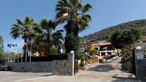 Ein Eingang mit Steinmauern, Flaggen und einem Gebäude mit der Aufschrift "KONI Village", sowie ein Berg im Hintergrund.
