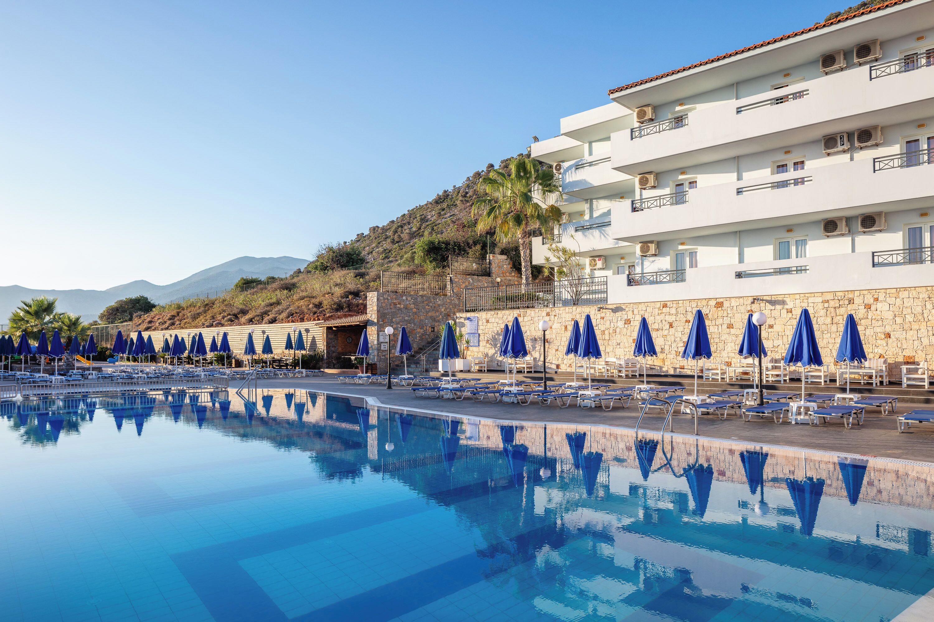 Ein Swimmingpool mit blauen Sonnenschirmen und Liegestühlen, ein weißes Gebäude mit Balkonen und eine Hügelkulisse im Hintergrund.
