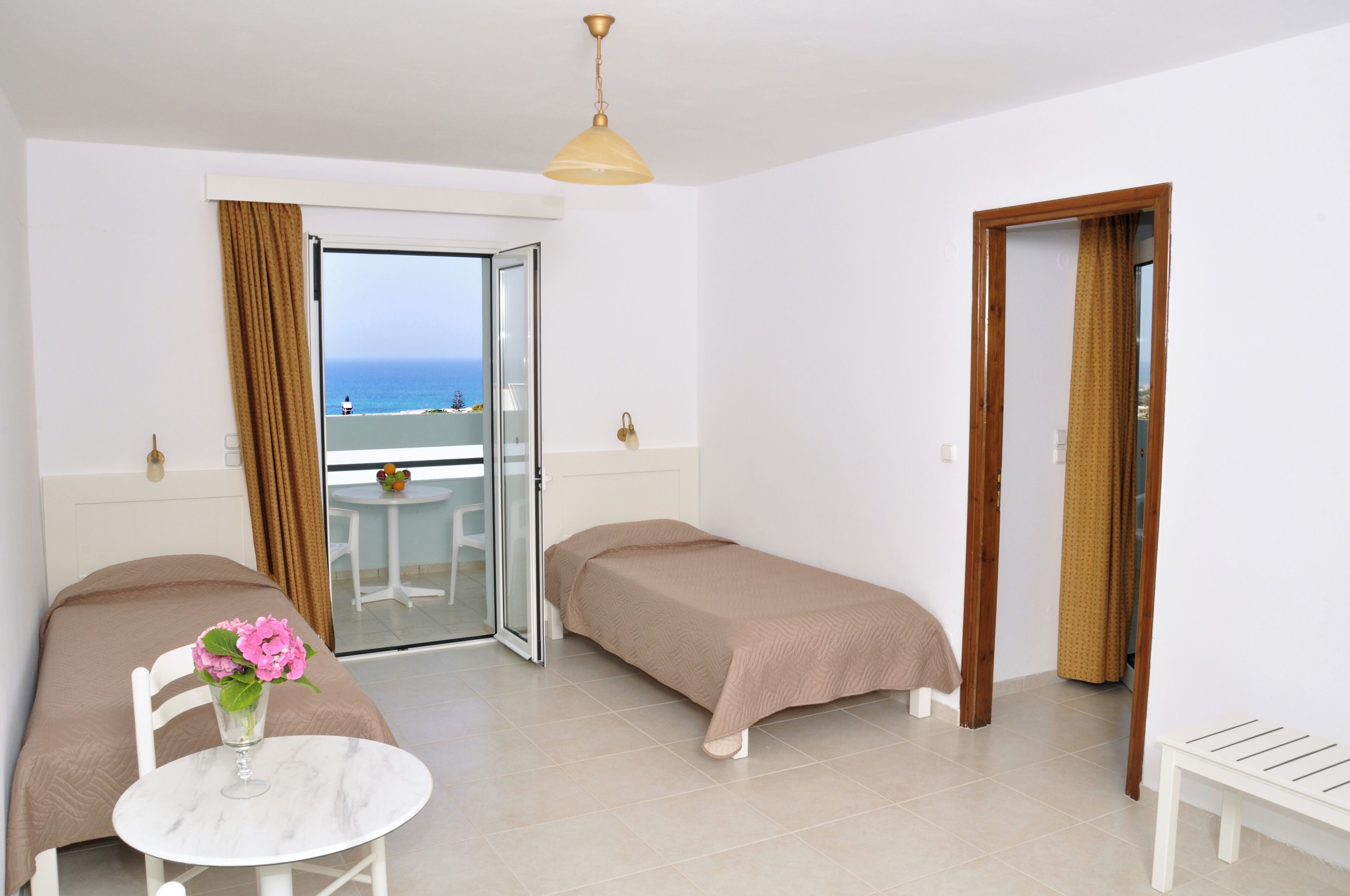 Quarto de hotel com duas camas de solteiro, uma mesinha com um vaso de flores, varanda com mesa e cadeiras e vista para o mar.