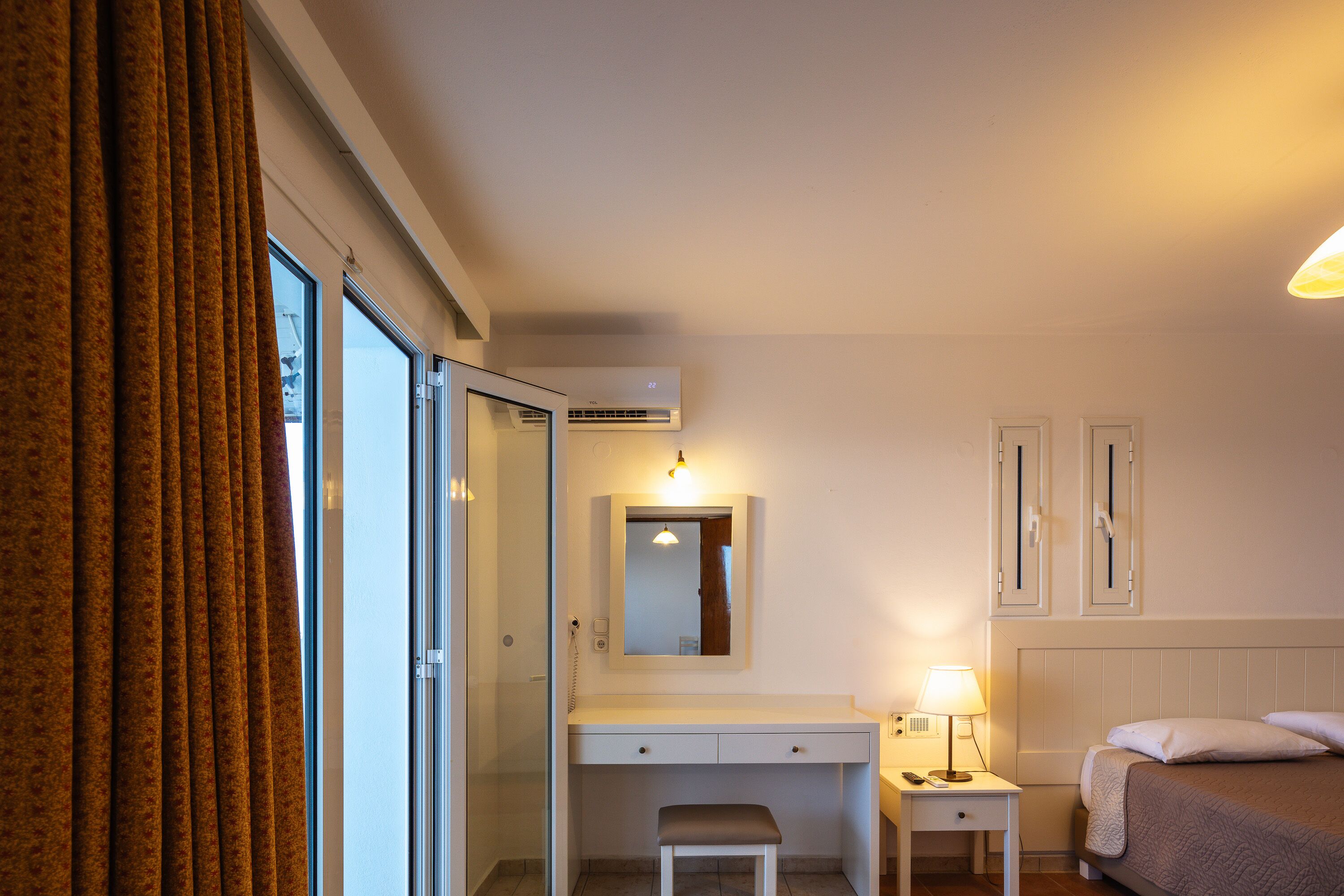 A hotel room with a bed, a desk, a chair, a bedside table with a lamp, a mirror, and a window with curtains.