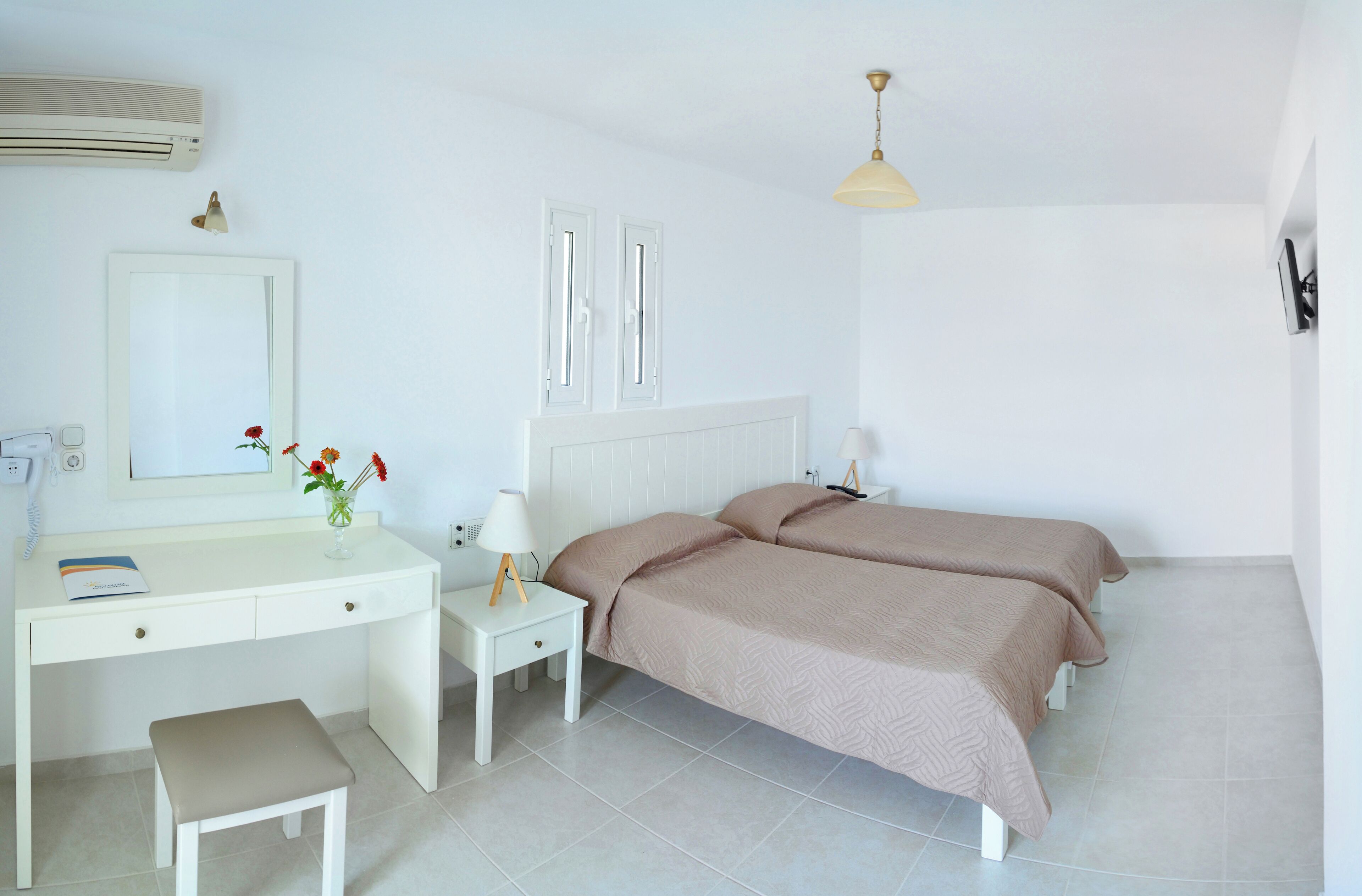 Une chambre avec un bureau blanc, une table de chevet avec une lampe, deux lits avec une literie beige, un miroir et un luminaire au plafond.
