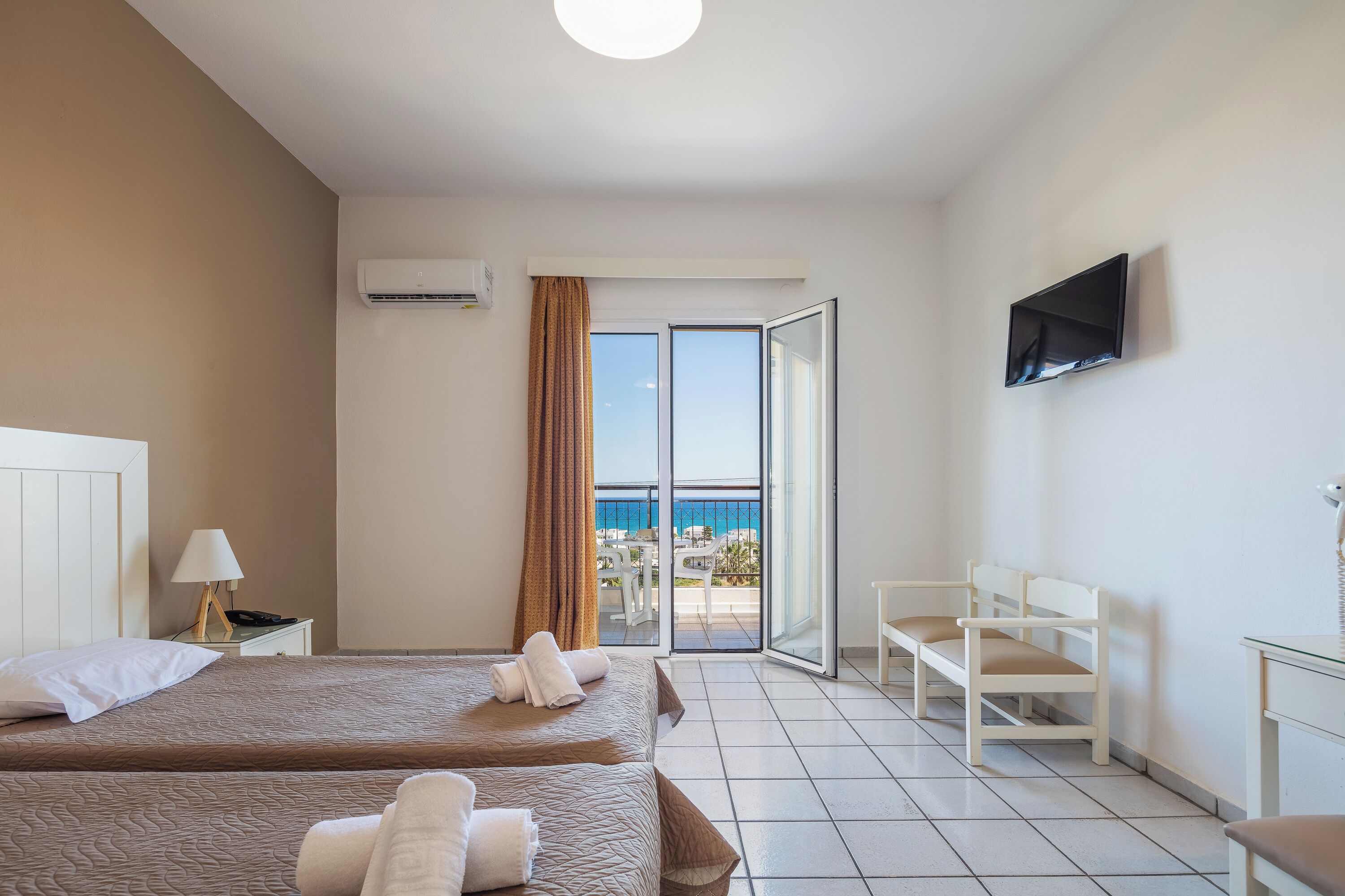 A hotel room with two beds, a TV, a desk, and a balcony with a view of the sea.