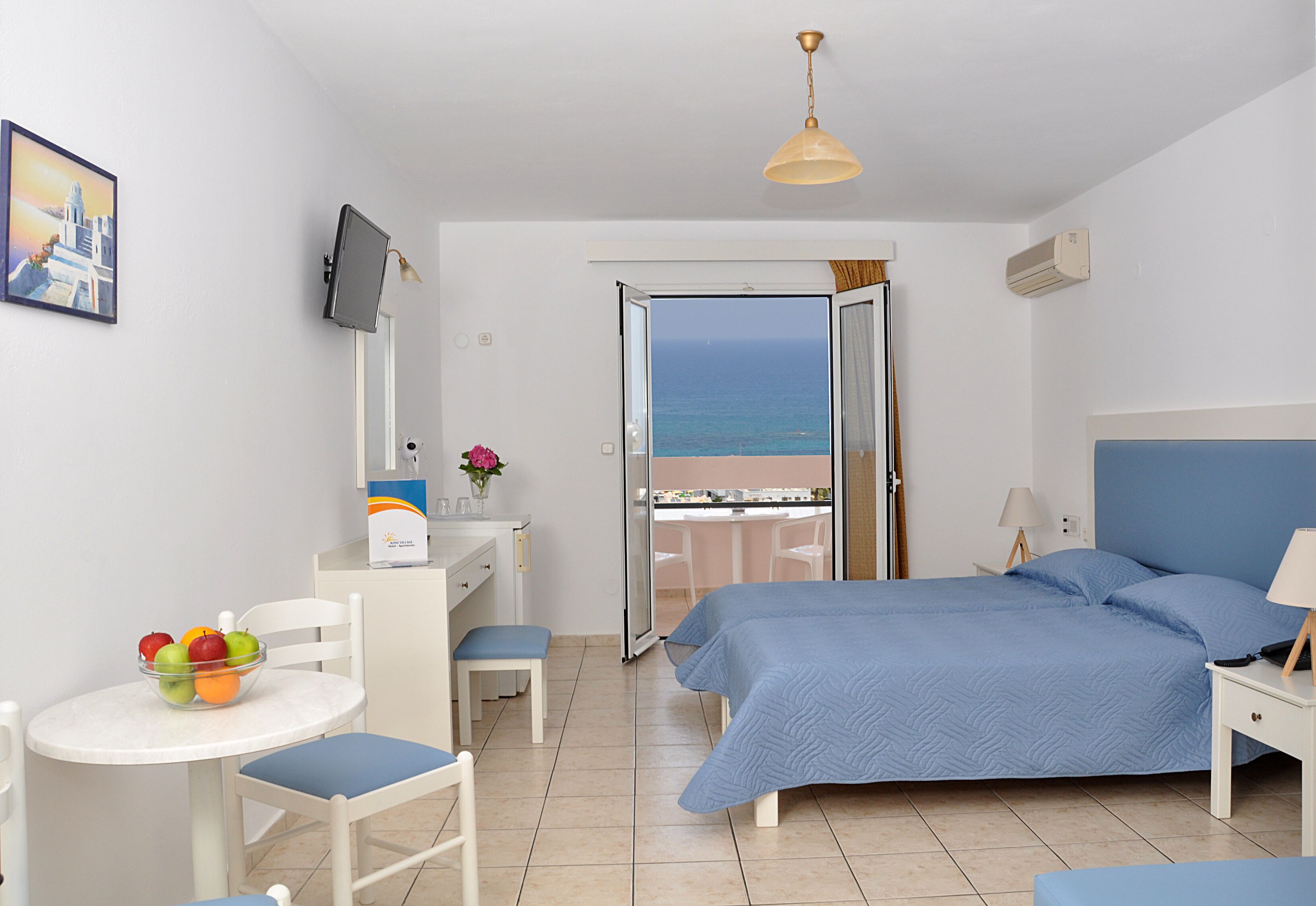 Habitación de hotel con cama, escritorio, silla, una mesita con fruta, televisor y vista al mar a través de una puerta abierta.