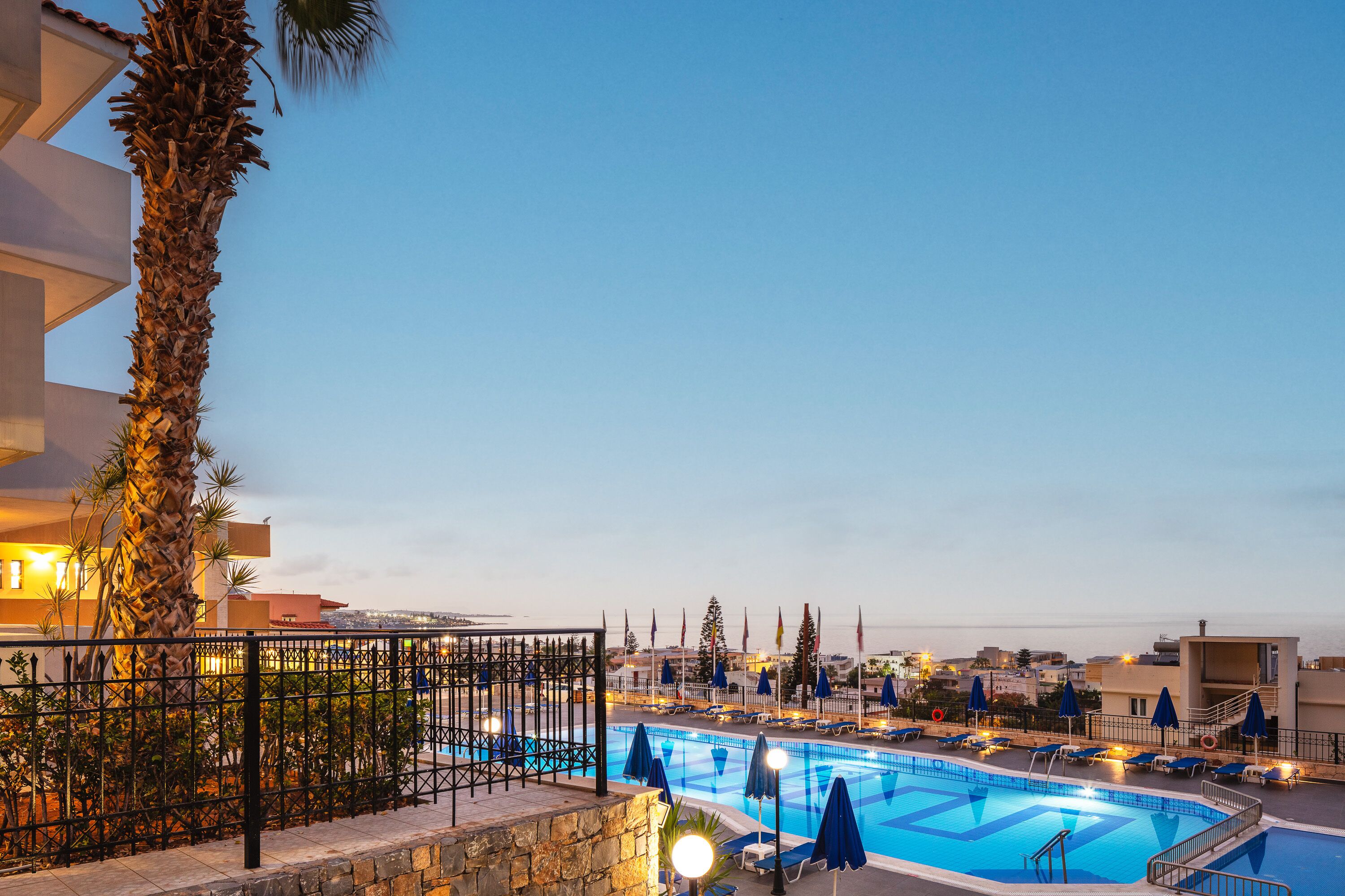A swimming pool with lounge chairs and umbrellas, surrounded by a stone wall and palm trees, with a view of the sea and buildings in the background.