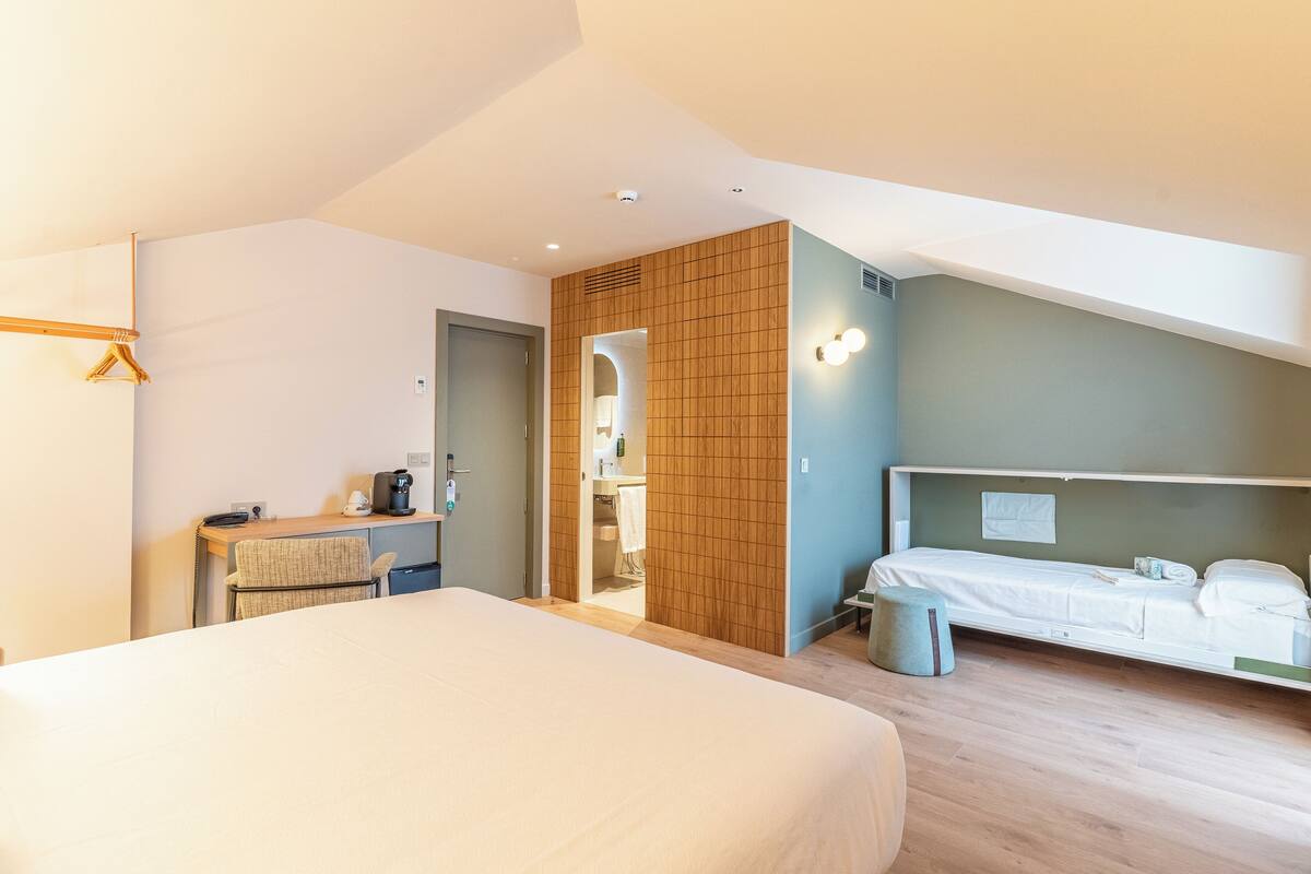 A modern hotel room with a large bed, a desk with a chair, a small stool, and a bathroom visible through an open door.