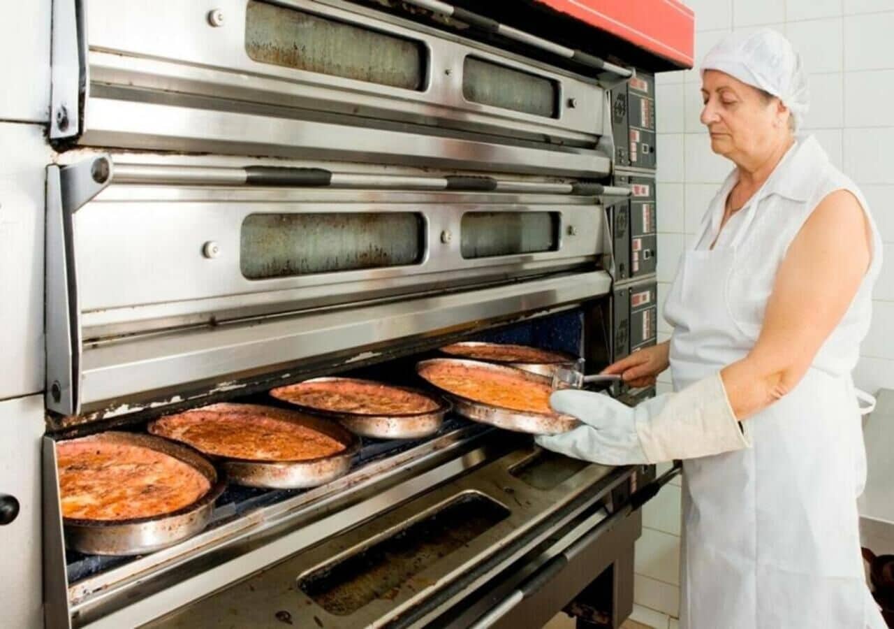 Una persona con uniforme blanco de chef operando un horno industrial con múltiples bandejas de comida.